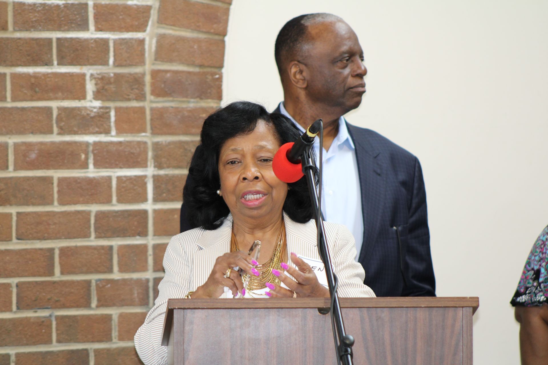 Woman speaking at podium, man standing behind her near brick wall.