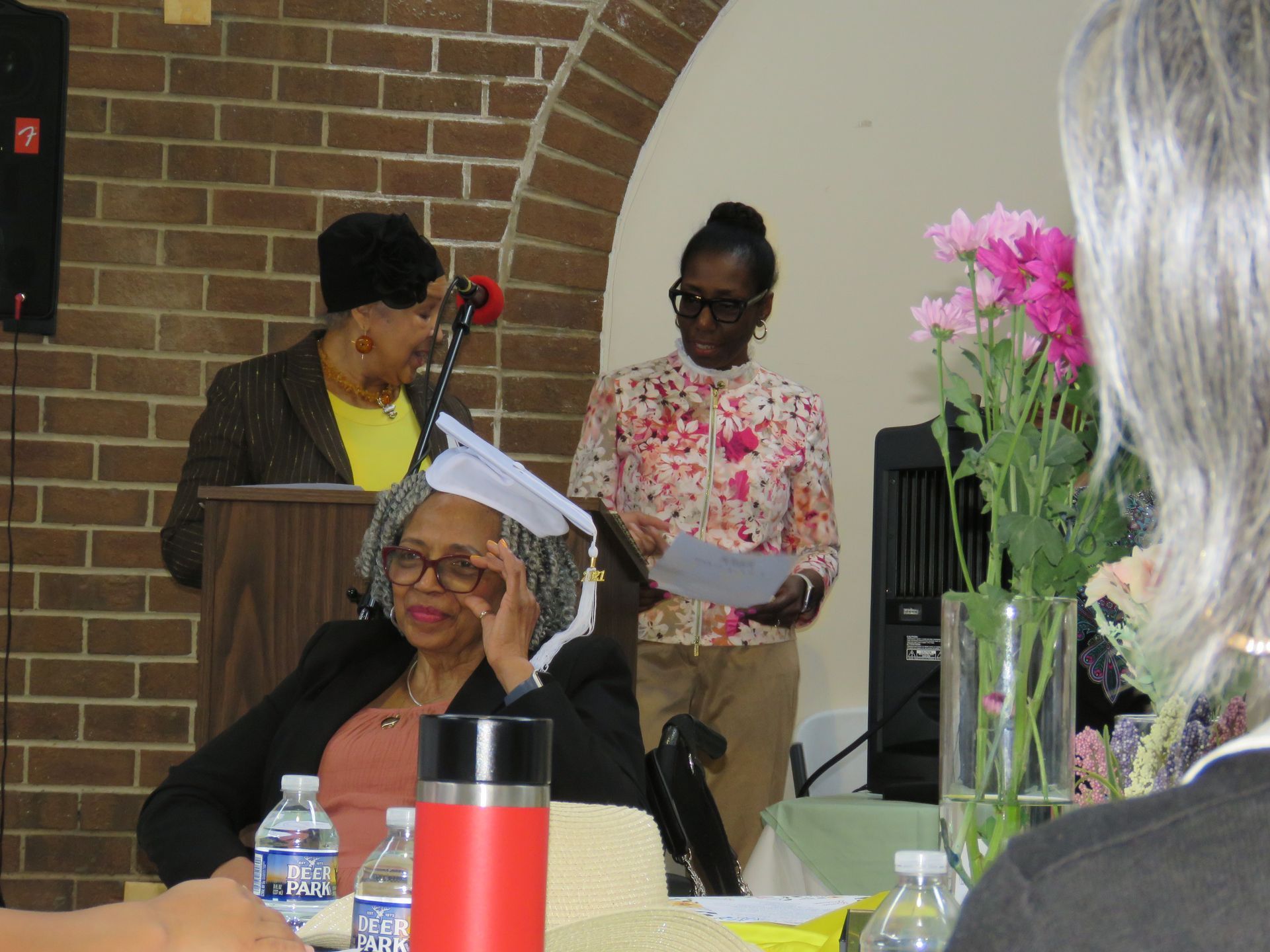 Three women at an event; one at a podium, one reading, one seated. Flowers and bottles on a table.