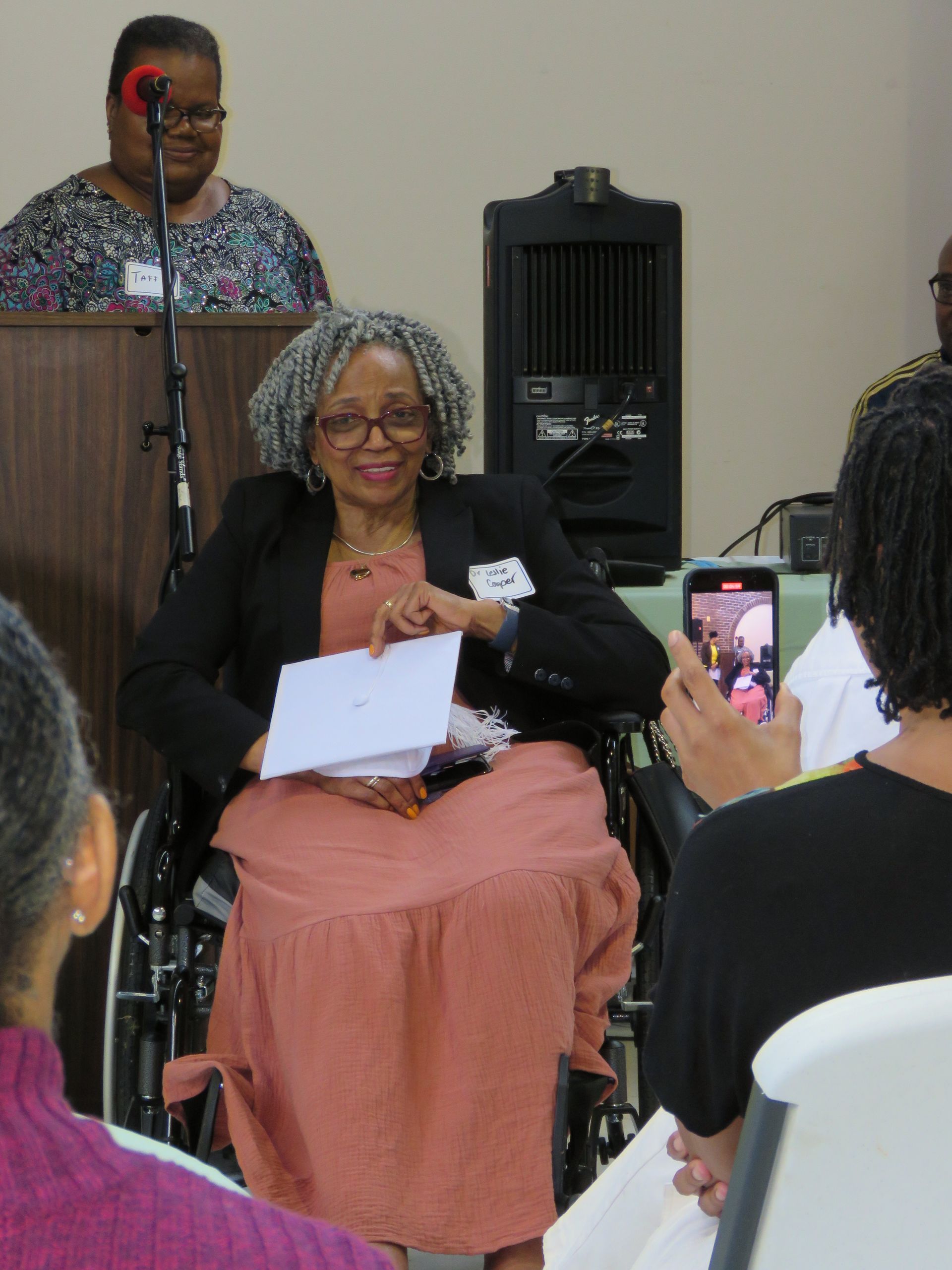 Woman in wheelchair smiling, holding papers at a podium. Another woman speaks at a podium behind her.