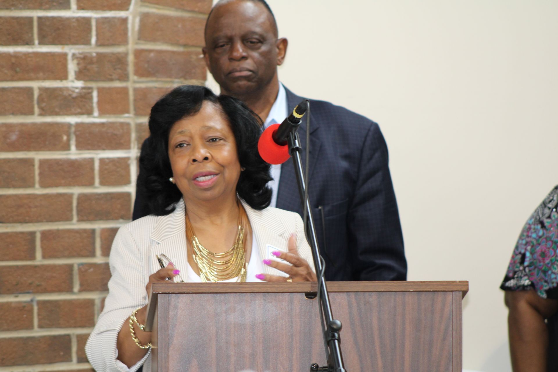 Woman speaking at a podium, man behind her, brick wall in the background.