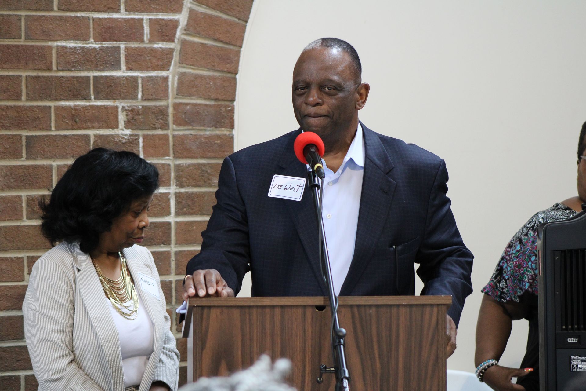 Man in a suit speaks at a podium; woman and man stand nearby. Brick wall in the background.