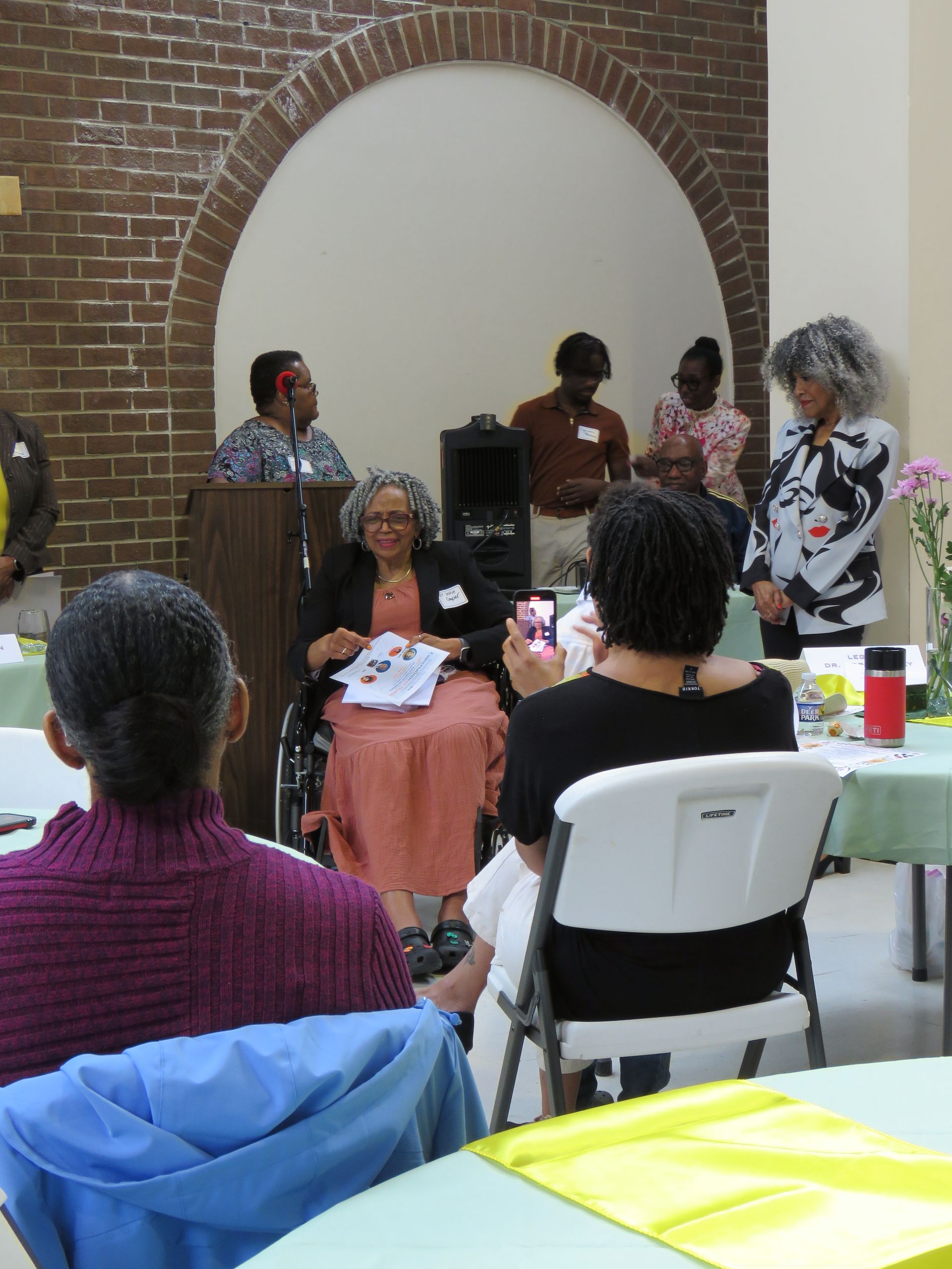 People at a gathering seated around tables; a speaker on a podium. Brick arch and neutral-toned wall as background.
