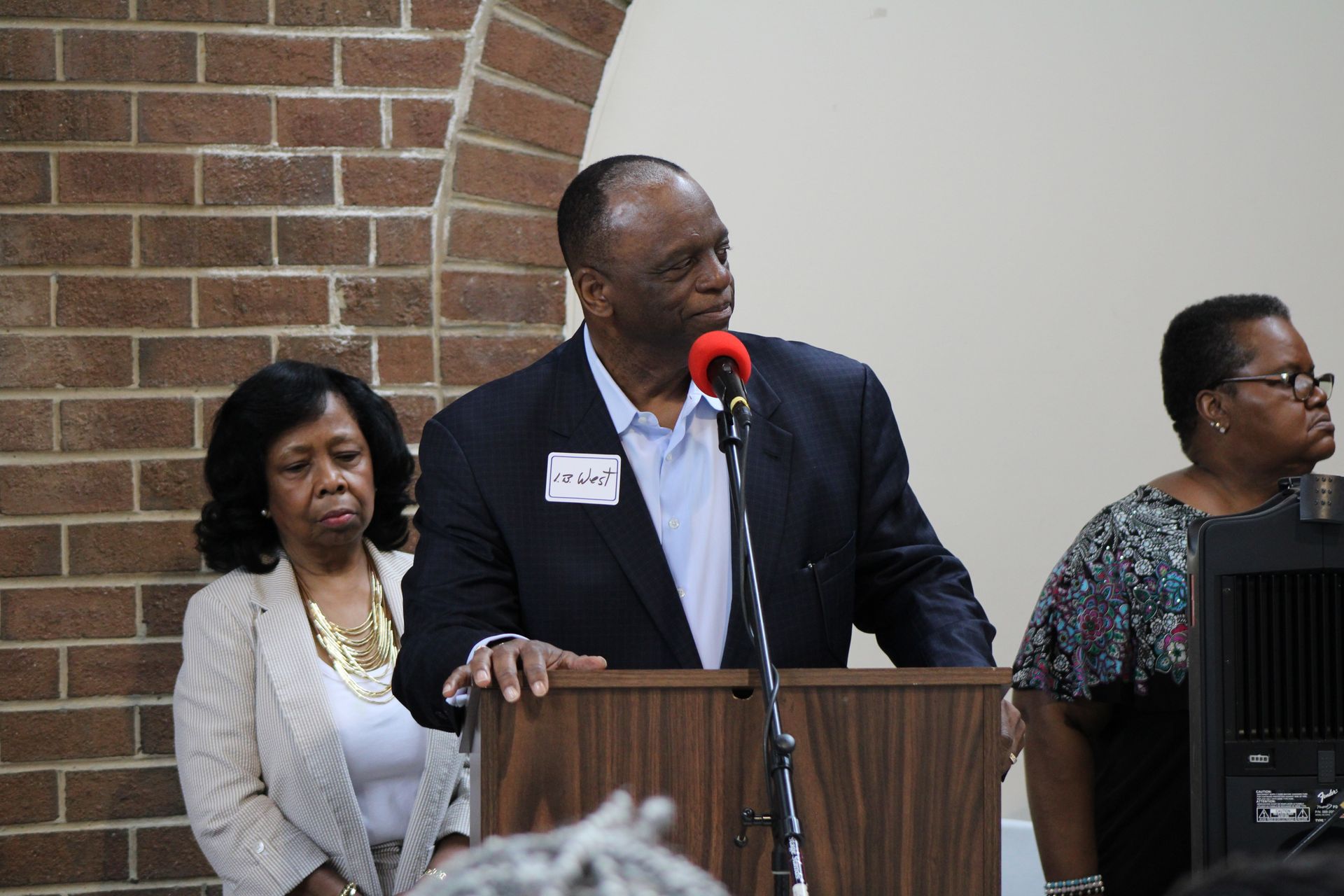 Man speaking at podium, two women standing beside him, brick wall background.