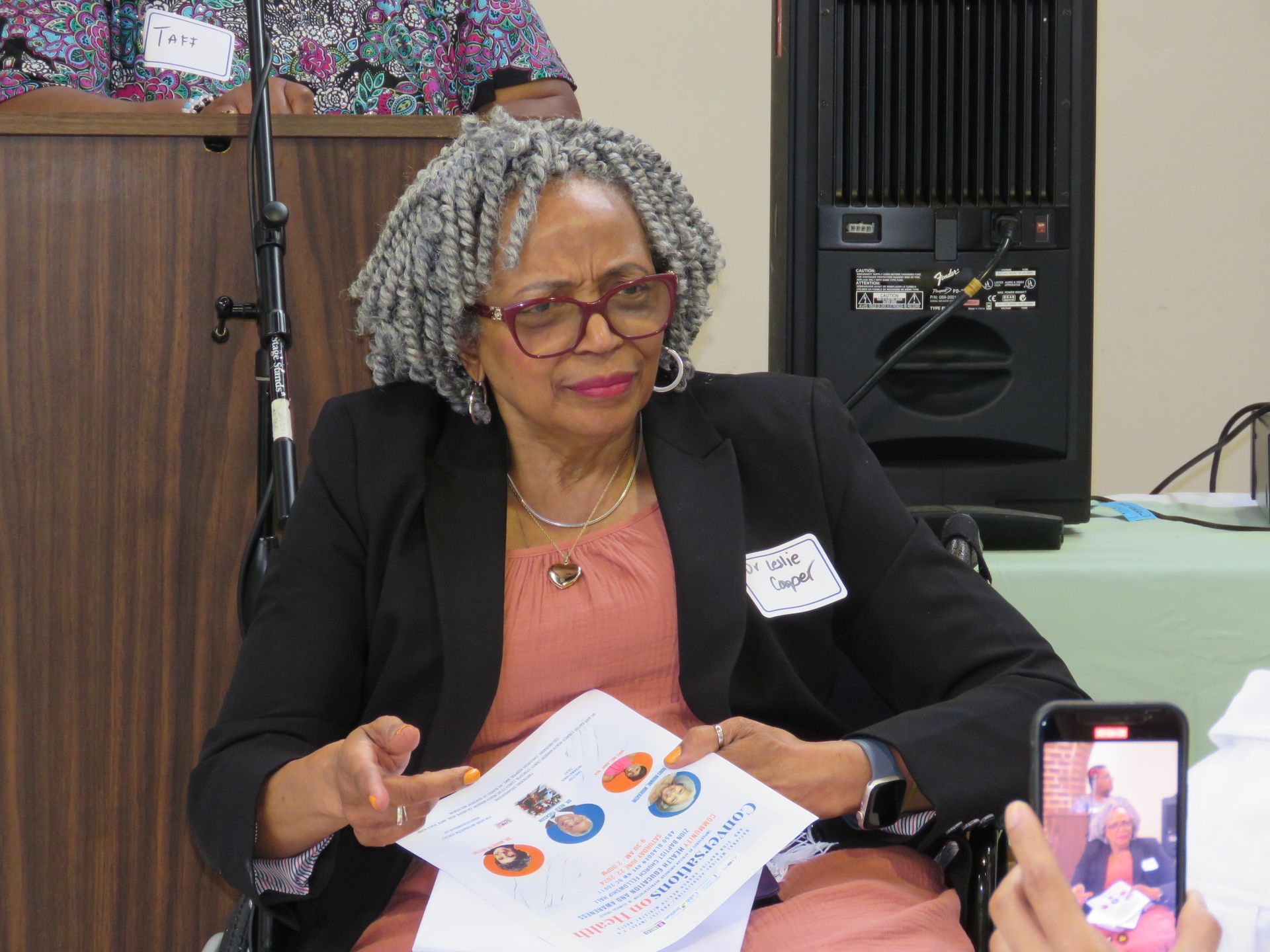 Woman in glasses holding paper, gesturing. Indoor setting, speaker podium in background.