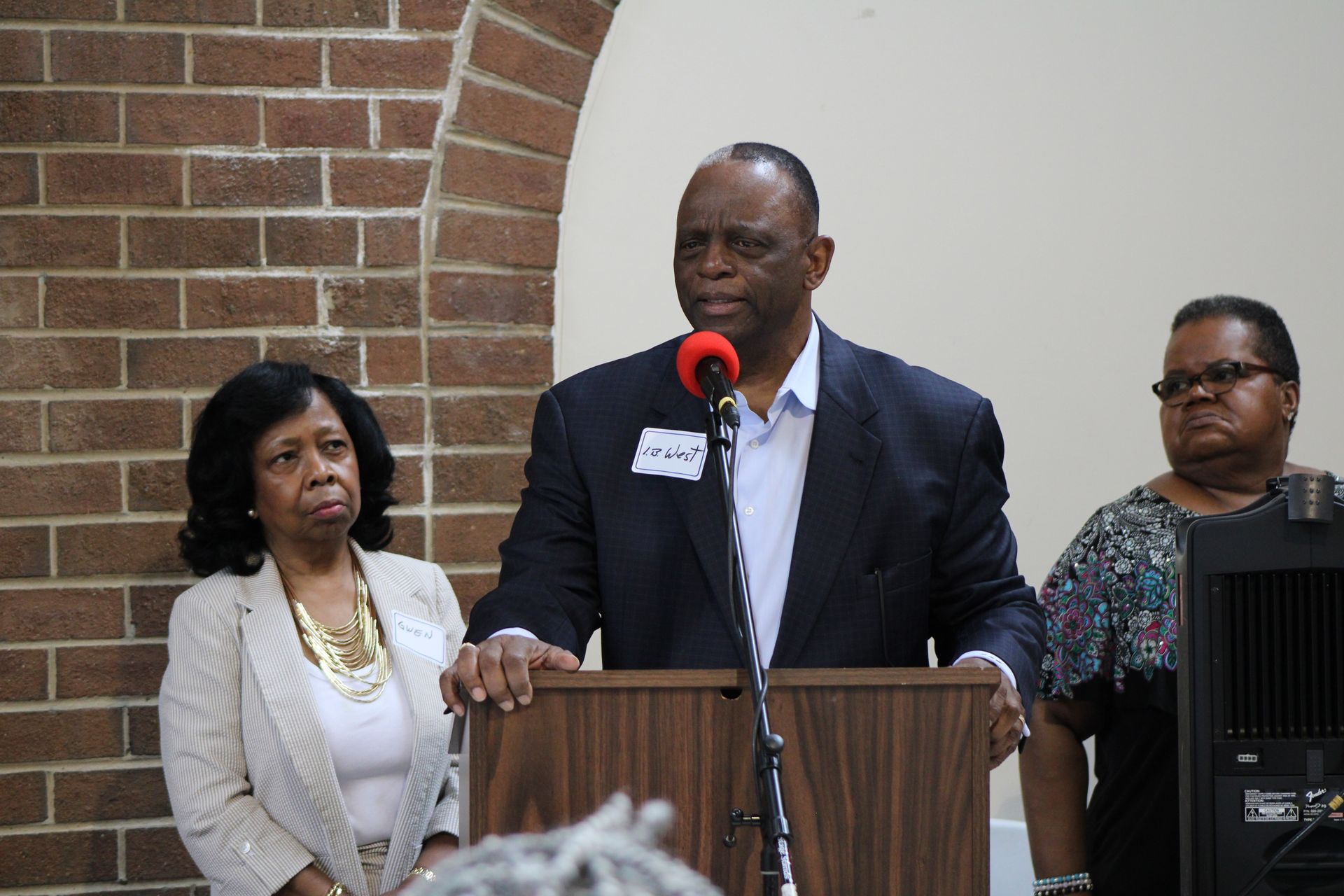 Man speaking at a podium with two women standing beside him. Brick wall backdrop.