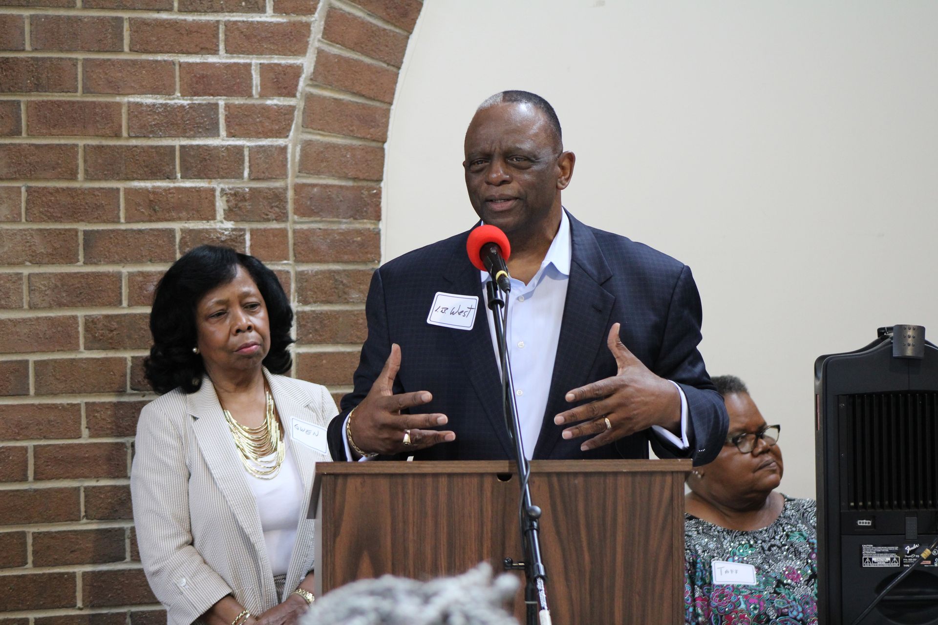 Man speaking at a podium, gesturing with hands; woman stands beside him. Brick wall background.