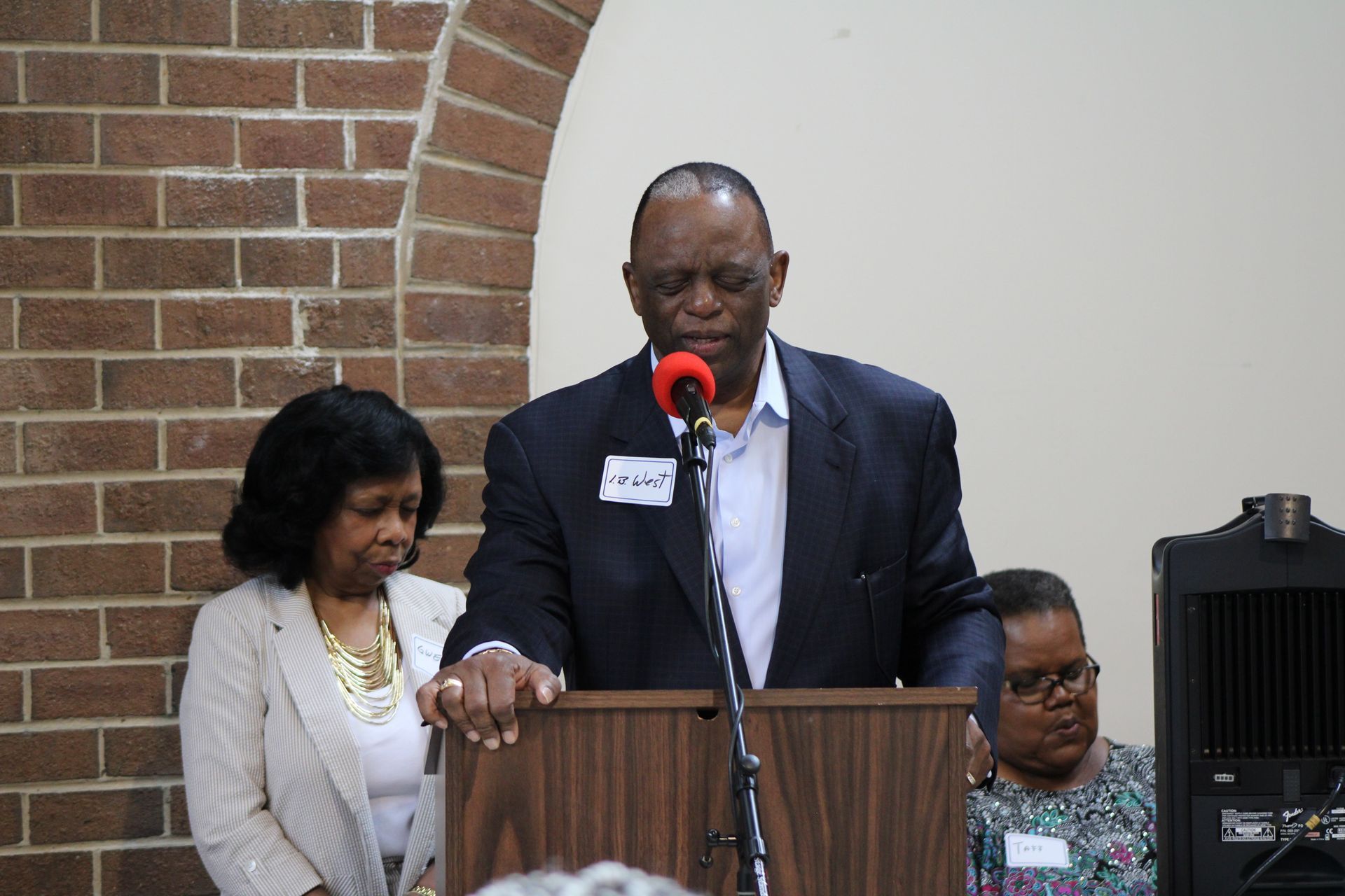 Man speaking at a podium with two women beside him. Brick wall backdrop.
