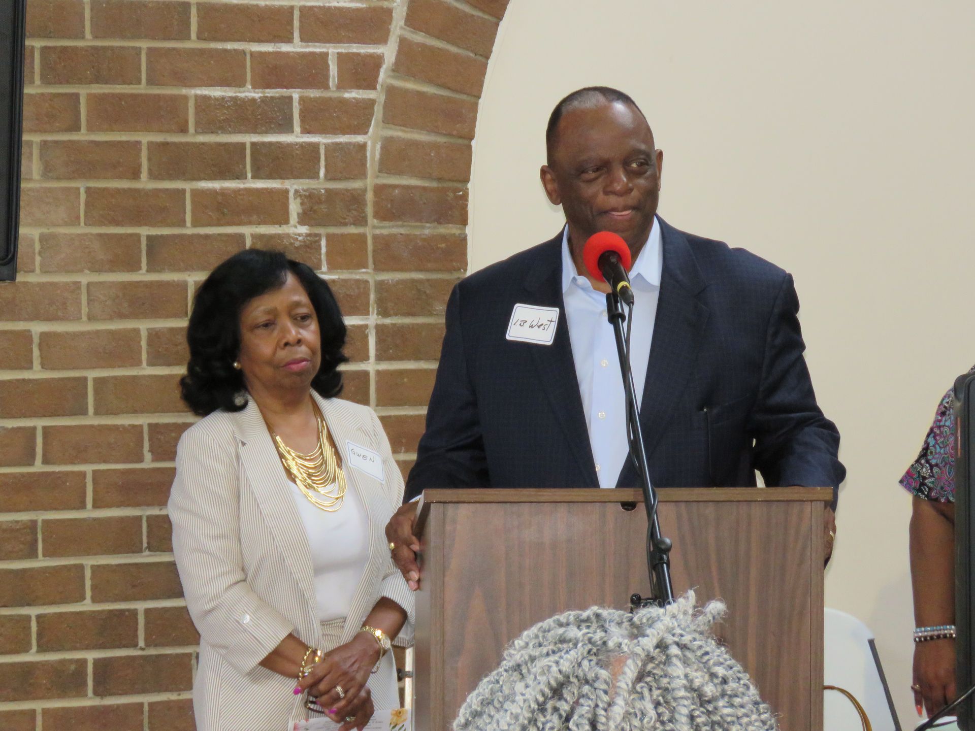 Man speaking at a podium, woman beside him, brick wall background.