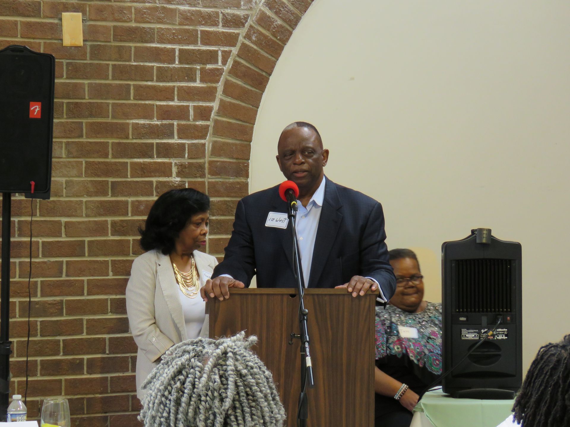 Man speaking at podium, two women beside him, brick wall background.