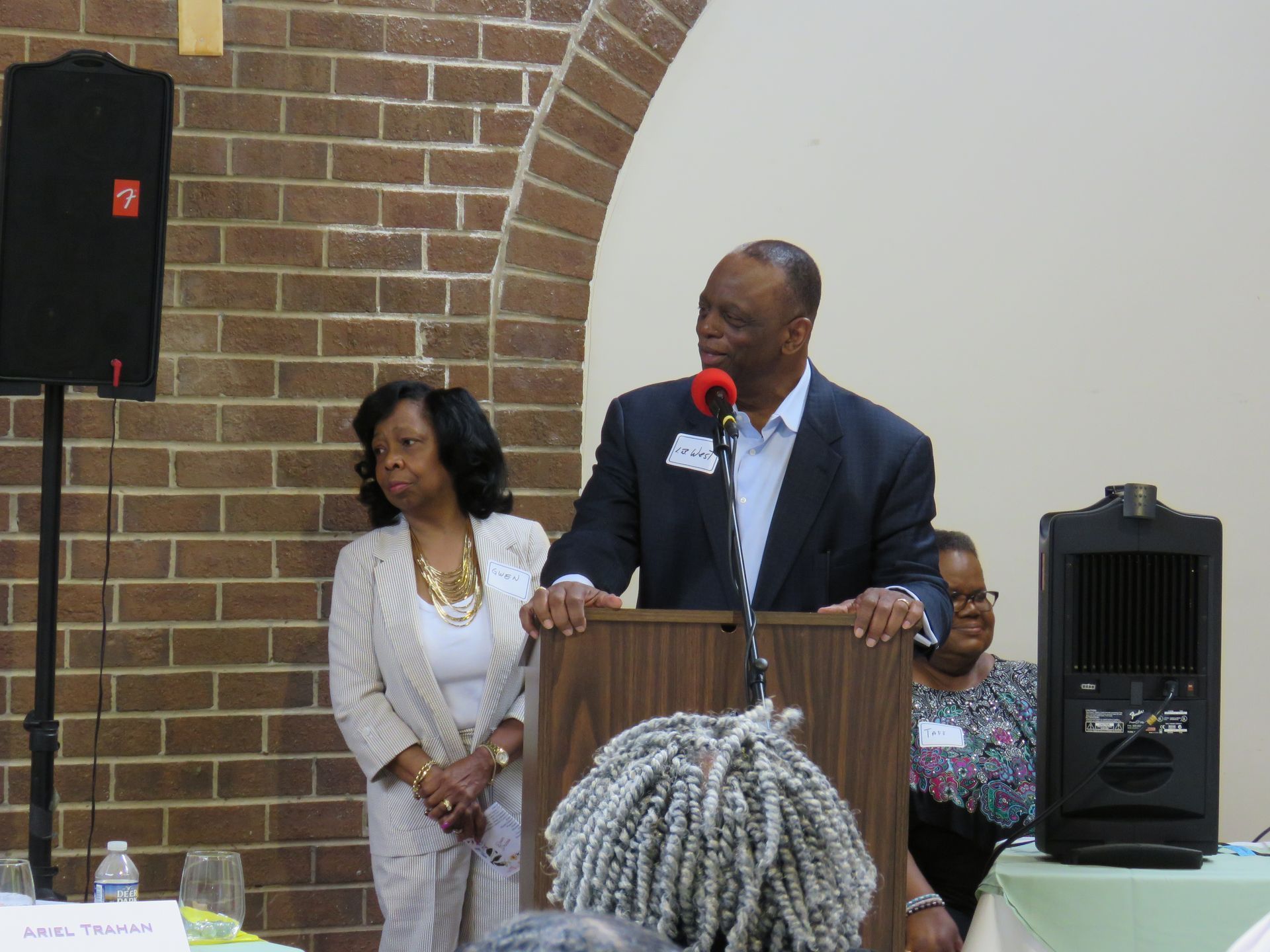 Man speaking at a podium with woman beside him; indoors, brick wall background.