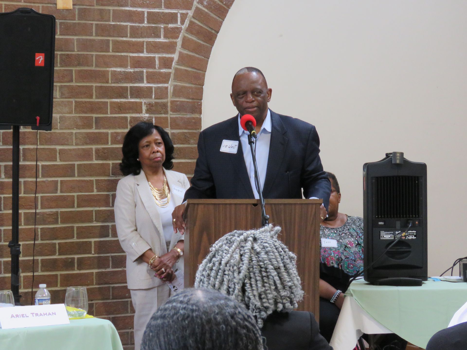 Man speaking at a podium with a microphone. Woman stands to his left. Brick wall background.