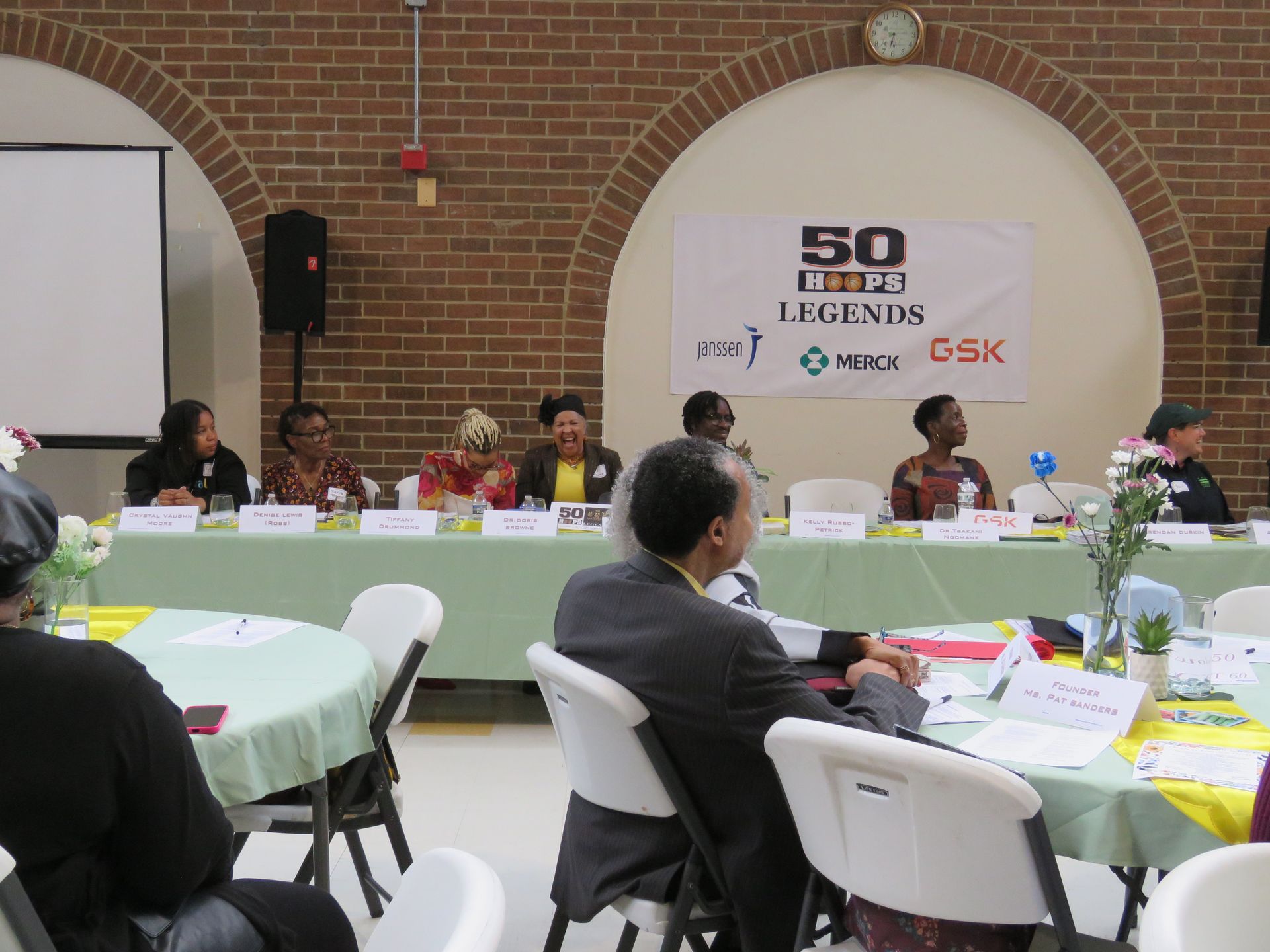 Panel discussion at a community event, people seated behind a table, audience at round tables. Banner reads 