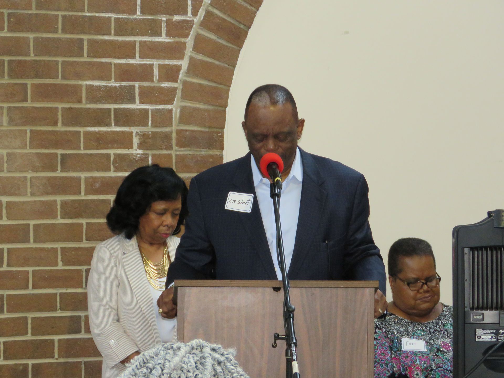 Man speaking at a podium, two women beside him, brick wall background.