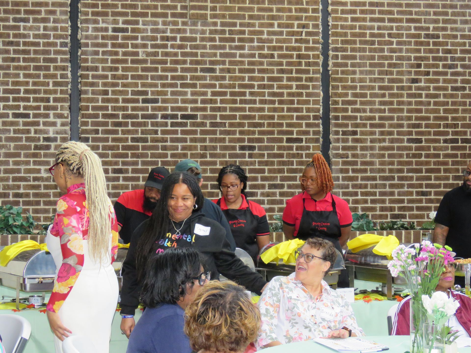 People at a food service area with brick wall background. Some are serving and others are seated.