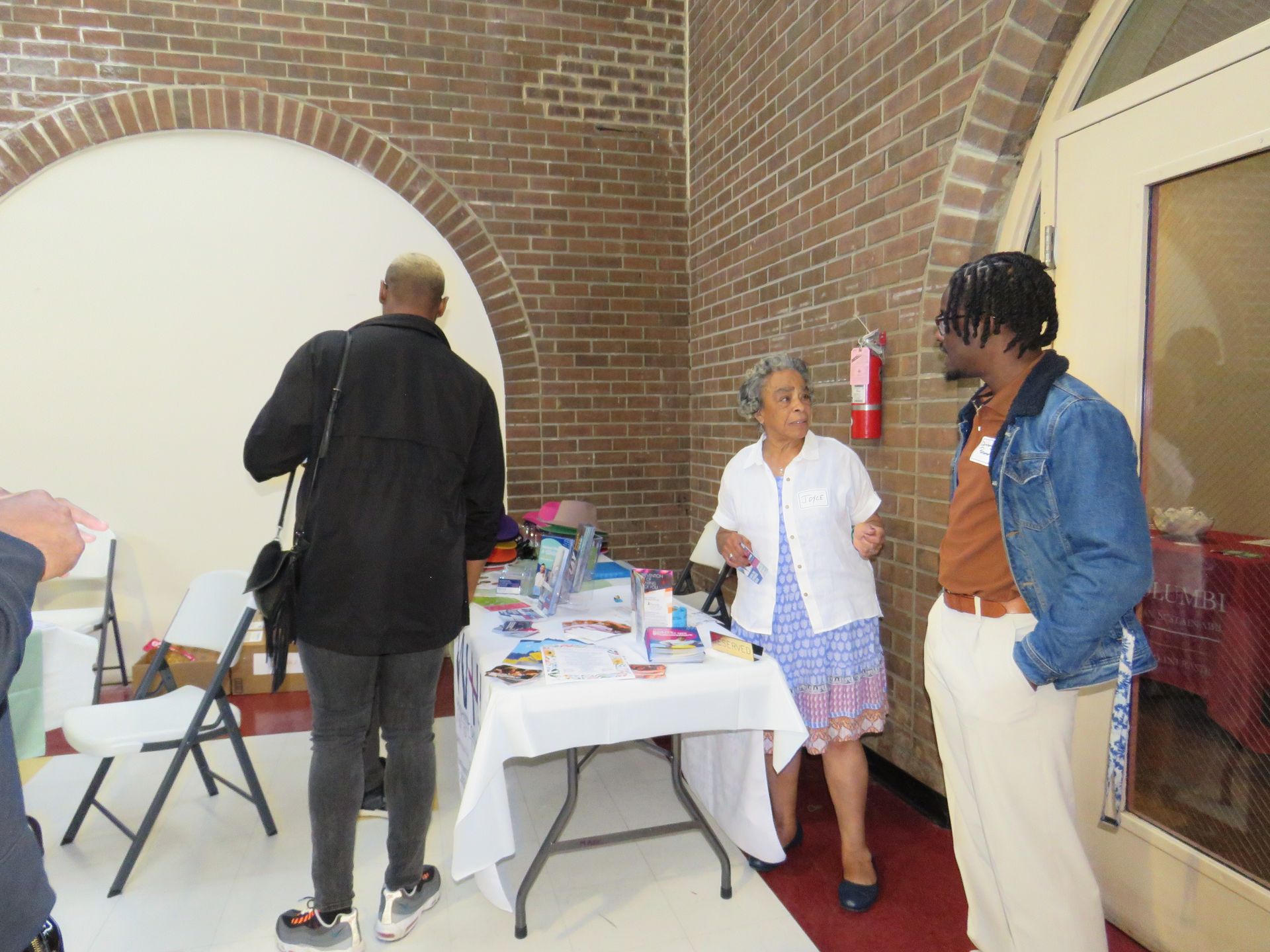 People at a table inside brick building. A person is handing items to someone else.