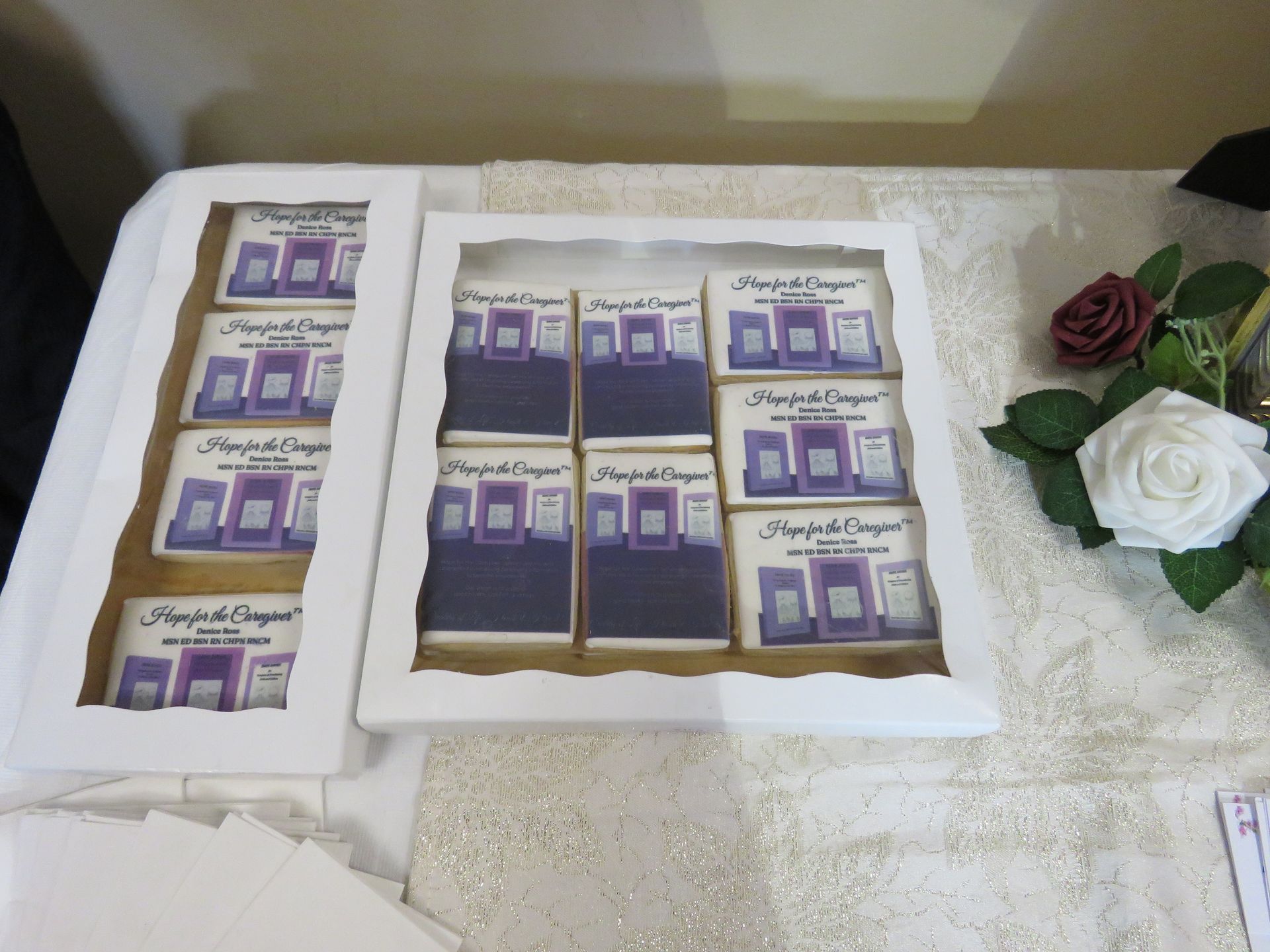 Cookies decorated with purple designs, in white boxes, on a table with flowers.