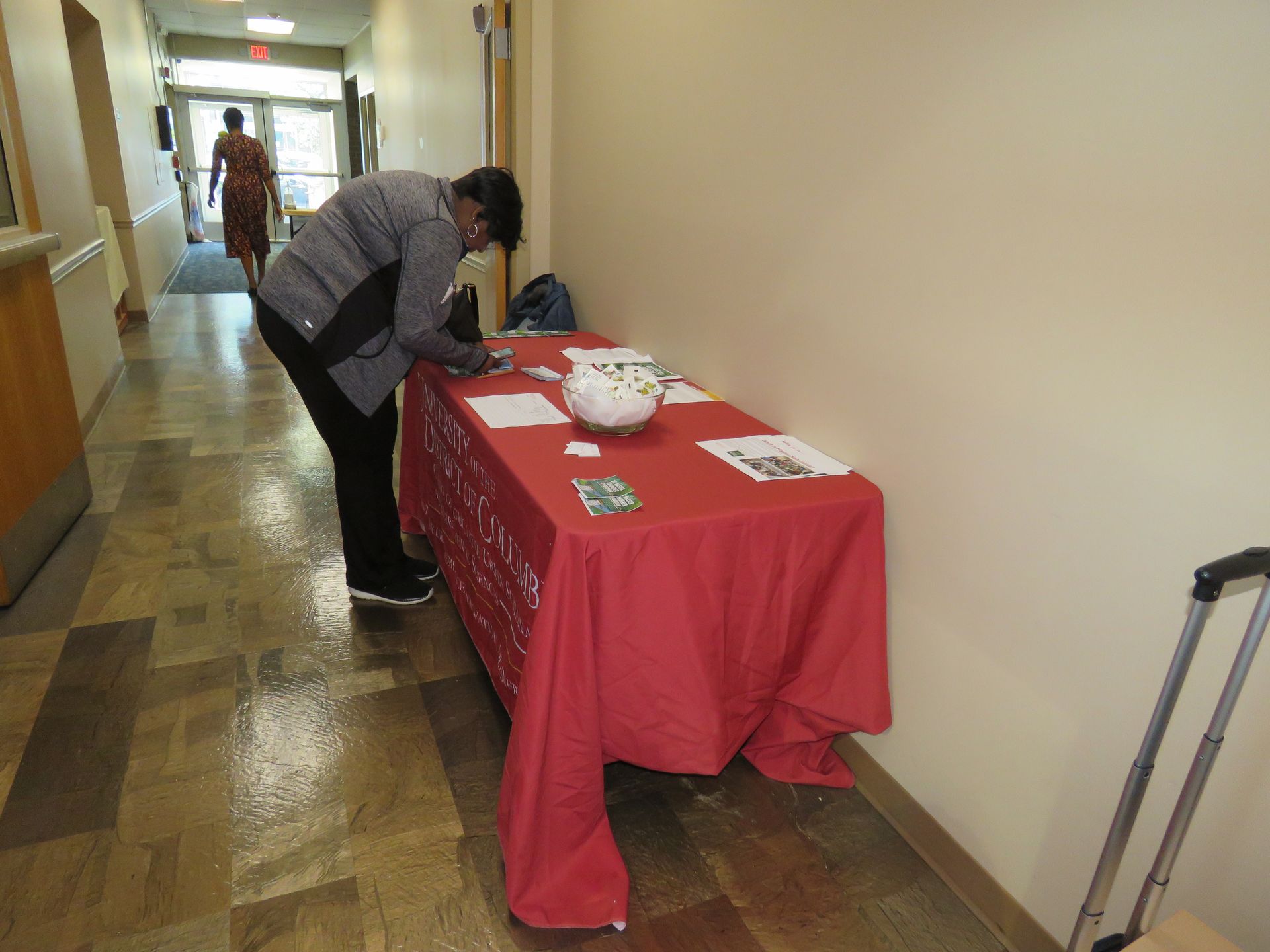 Person at a table with a red tablecloth in a hallway, writing. Another person walks in the background.