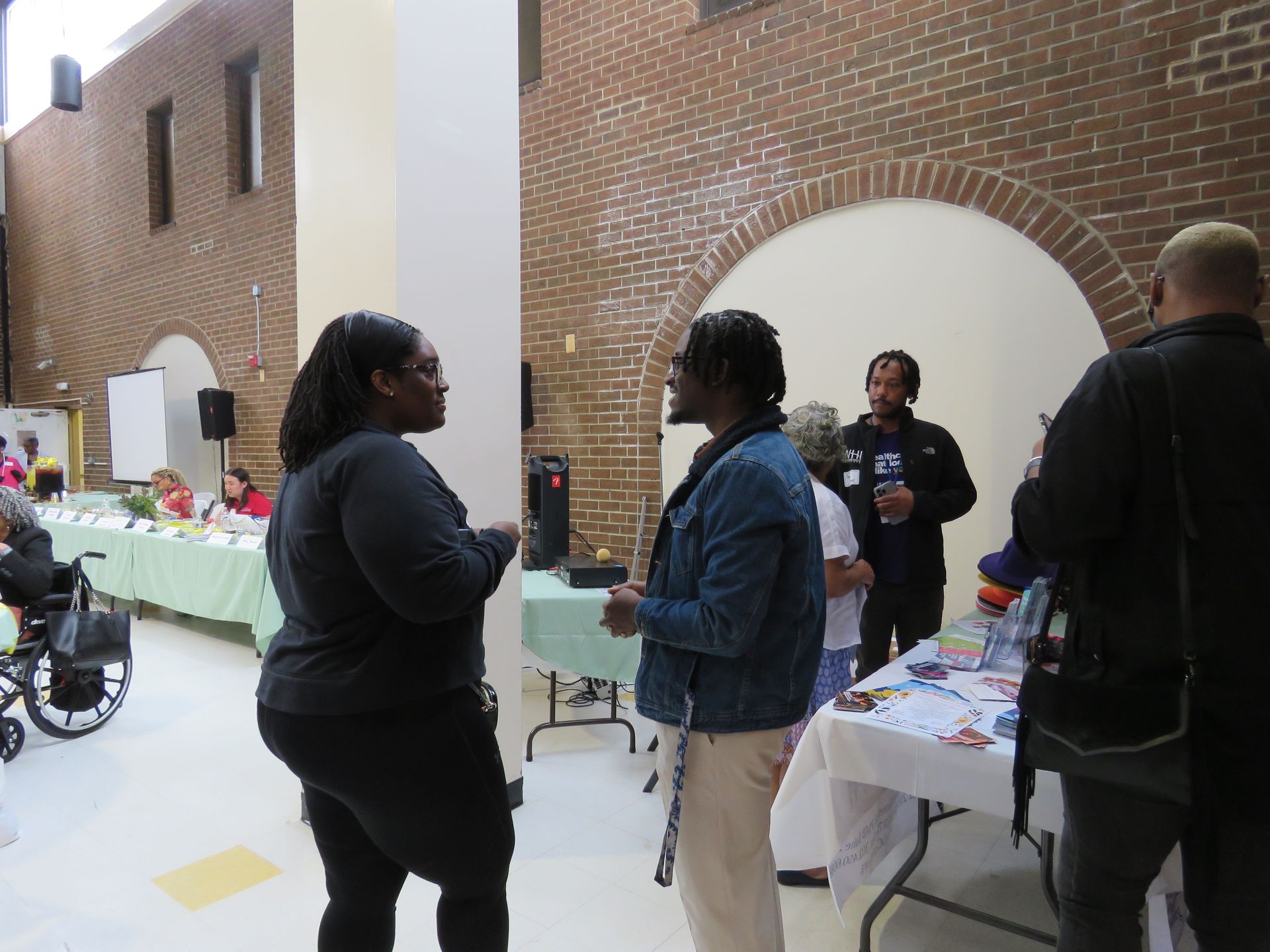People talking near tables in a brick-walled room. Some tables have items on them. One person is in a wheelchair.
