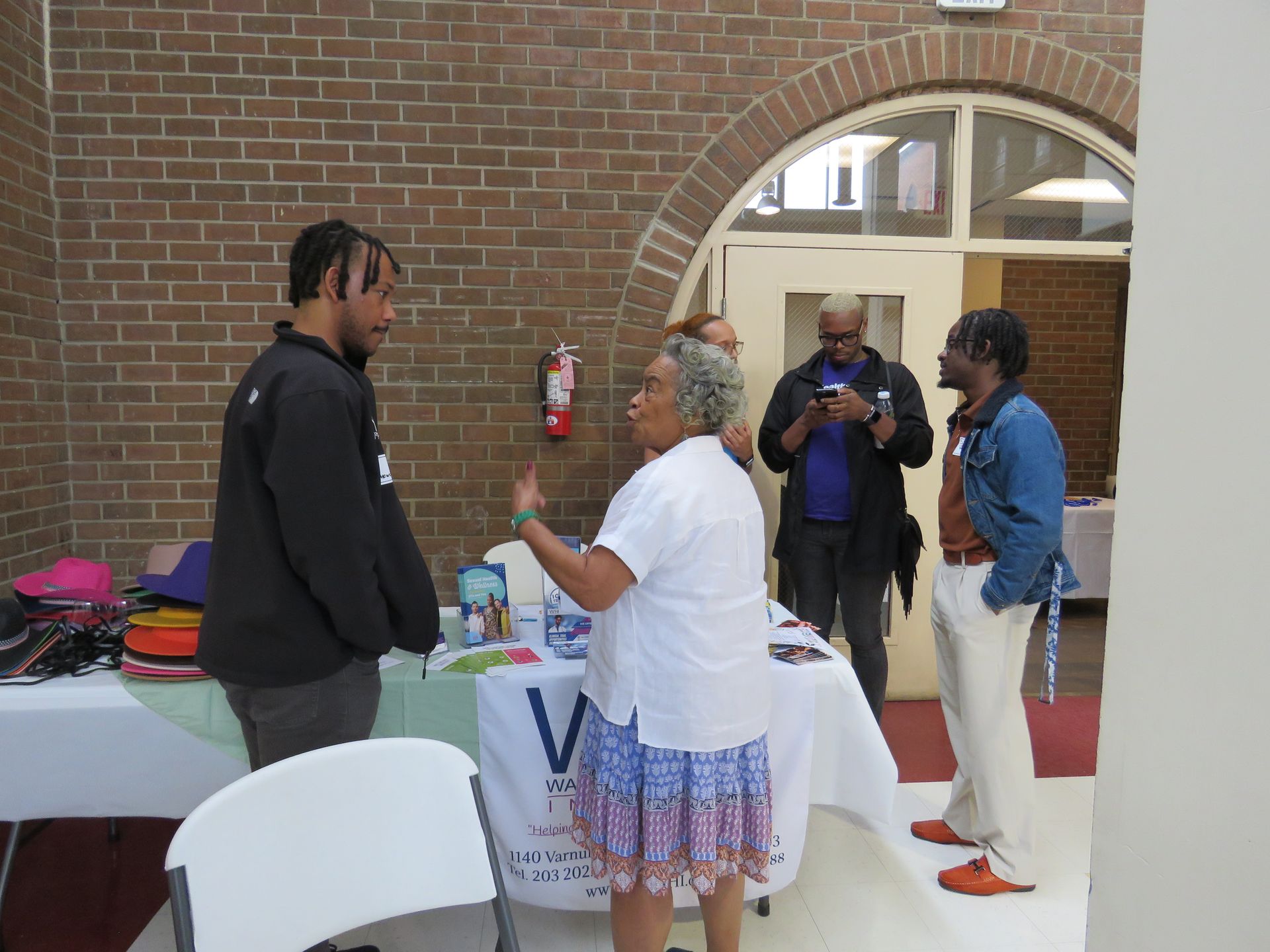 A woman gestures to a man at a table, two other men watch near a brick wall.
