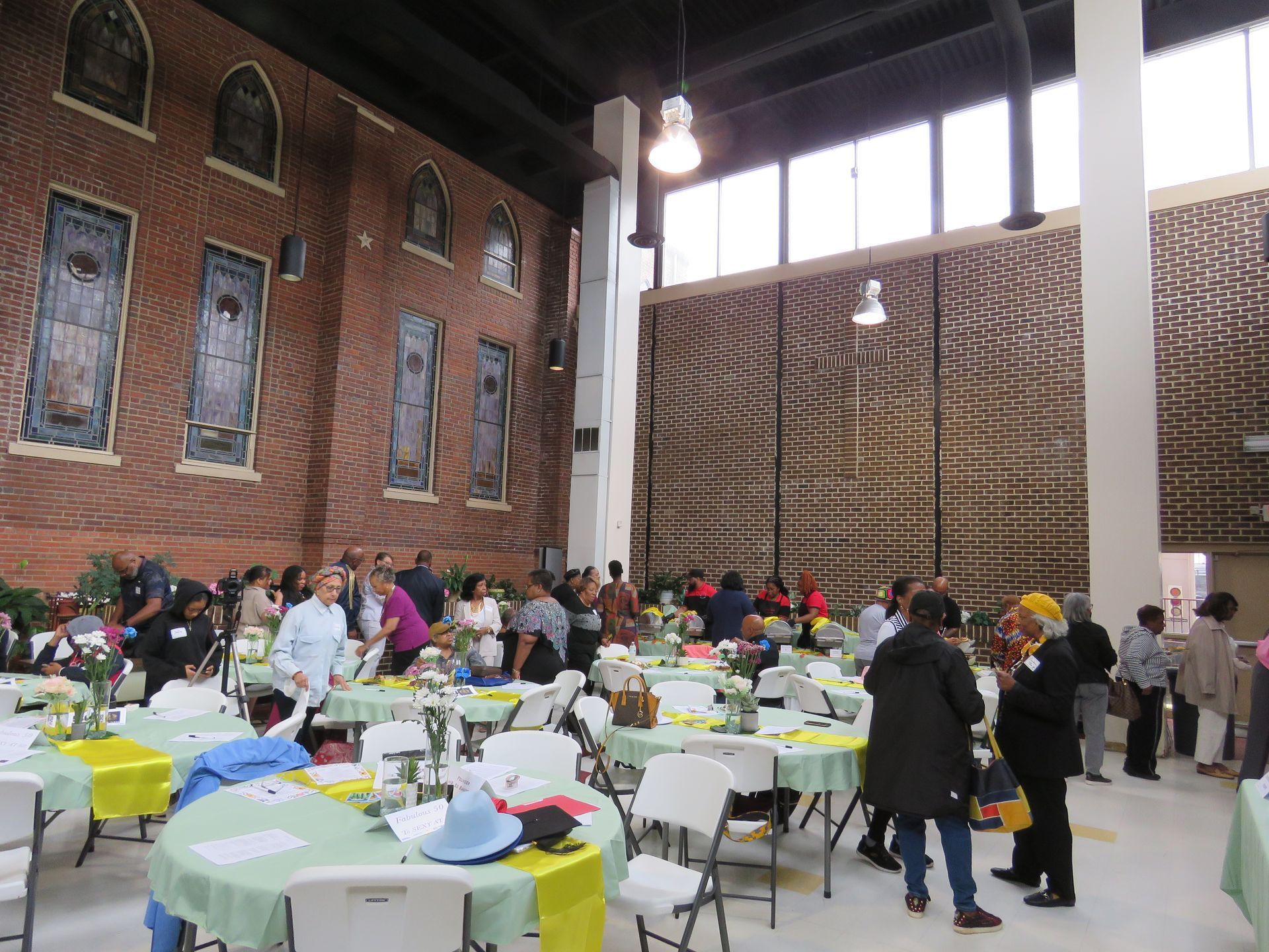 Large room with people at round tables; brick wall with stained glass, tall screen.