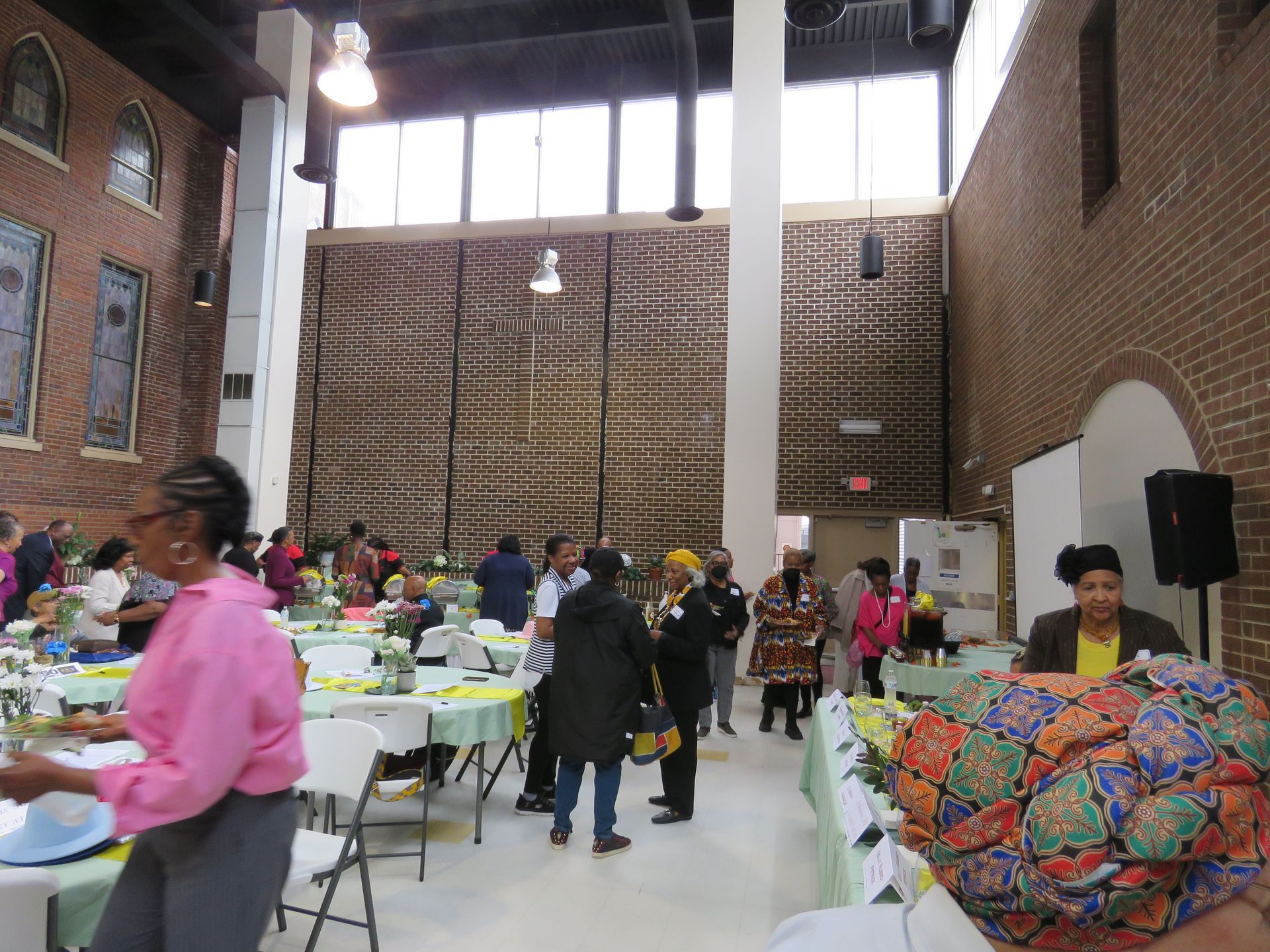 People gather in a large room with tables, some with food. Brick walls and high windows are visible.