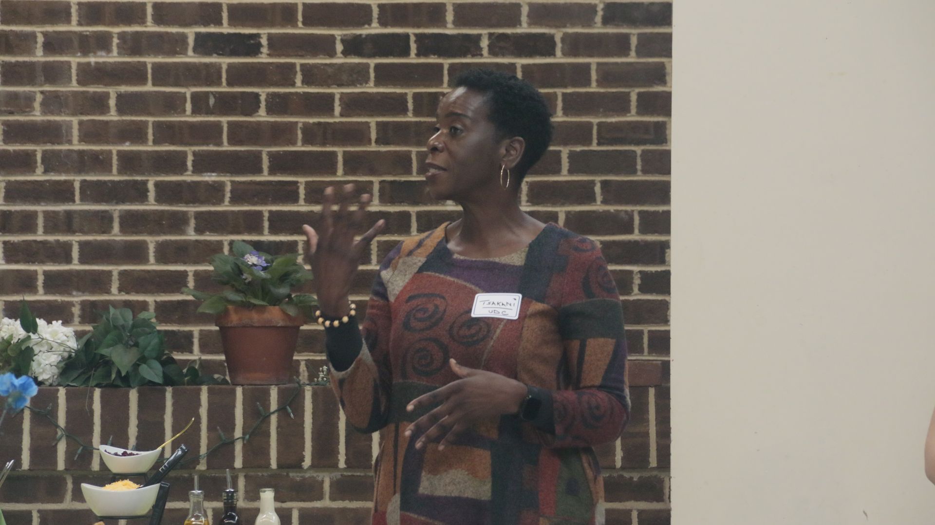 Woman speaking, gesturing with hands, in front of a brick wall and floral decorations.