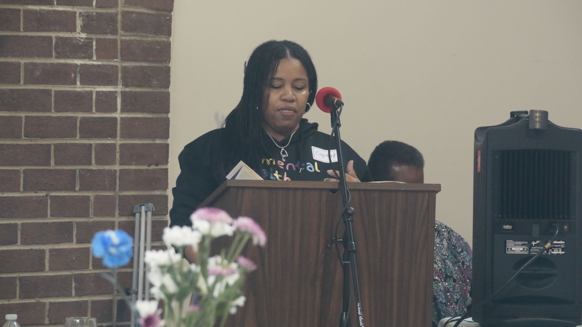 Woman speaking at a podium, microphone, flowers on table, brick wall background.