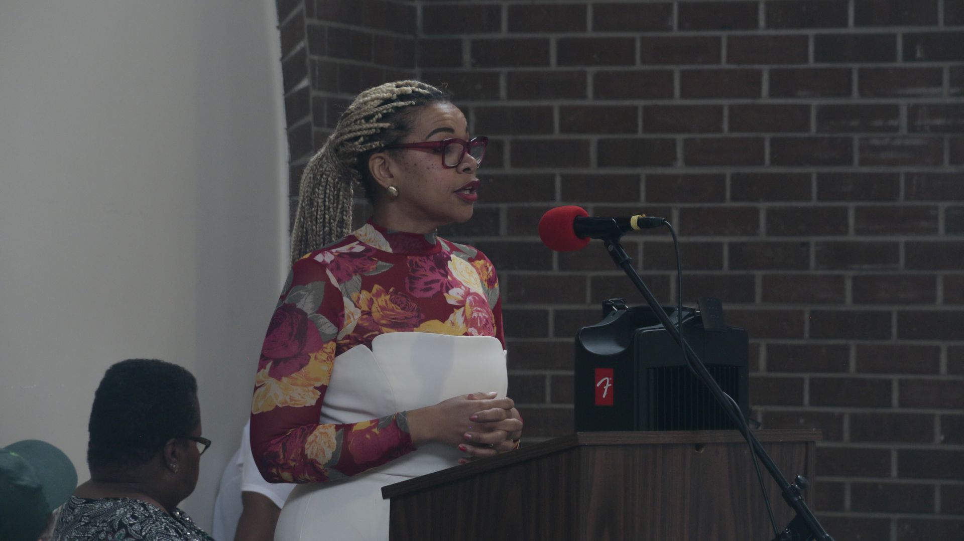 Woman speaking at a podium with a microphone in front of a brick wall.