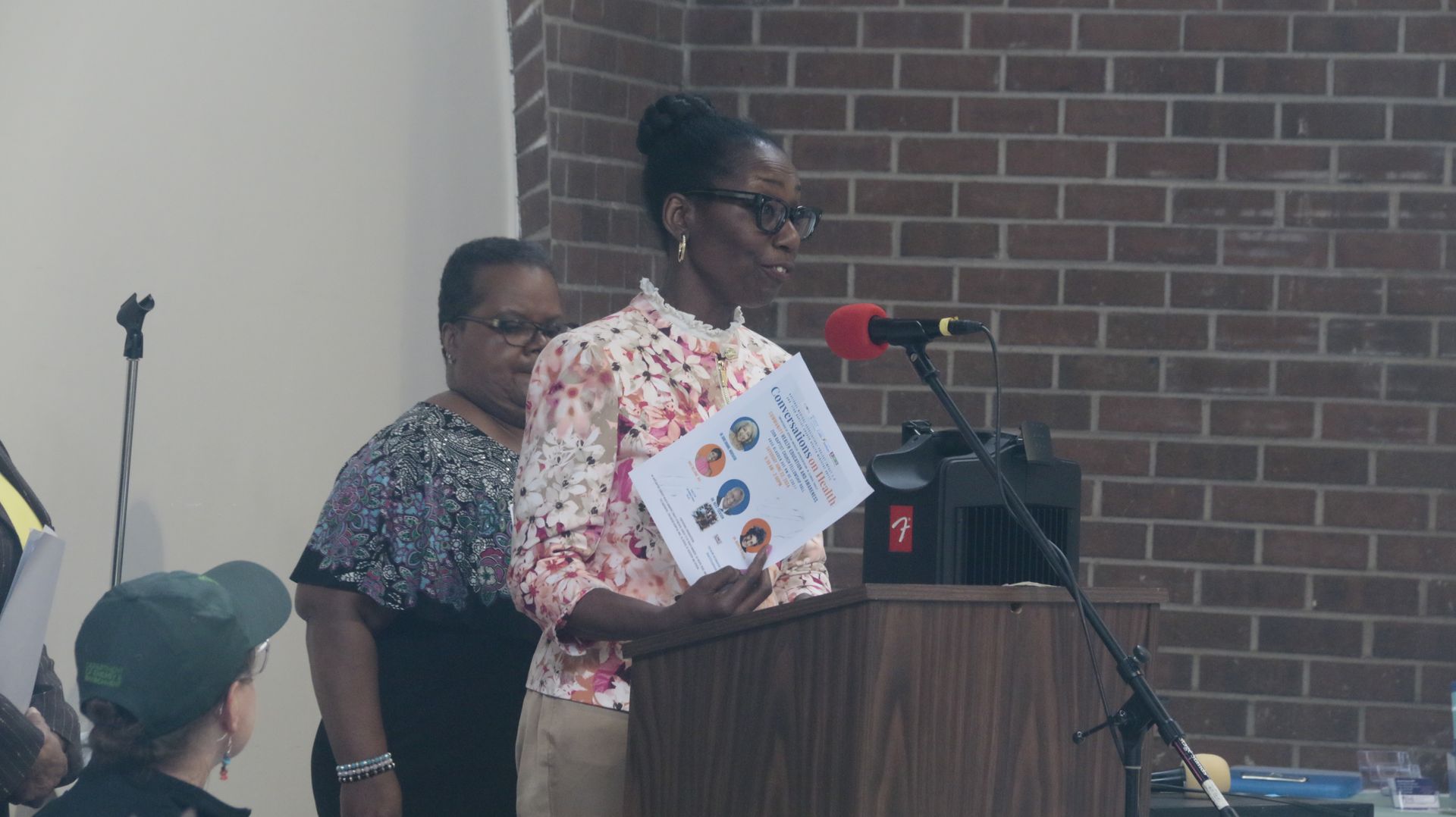 Woman speaking at a podium, holding papers, with two women standing behind her against a brick wall.