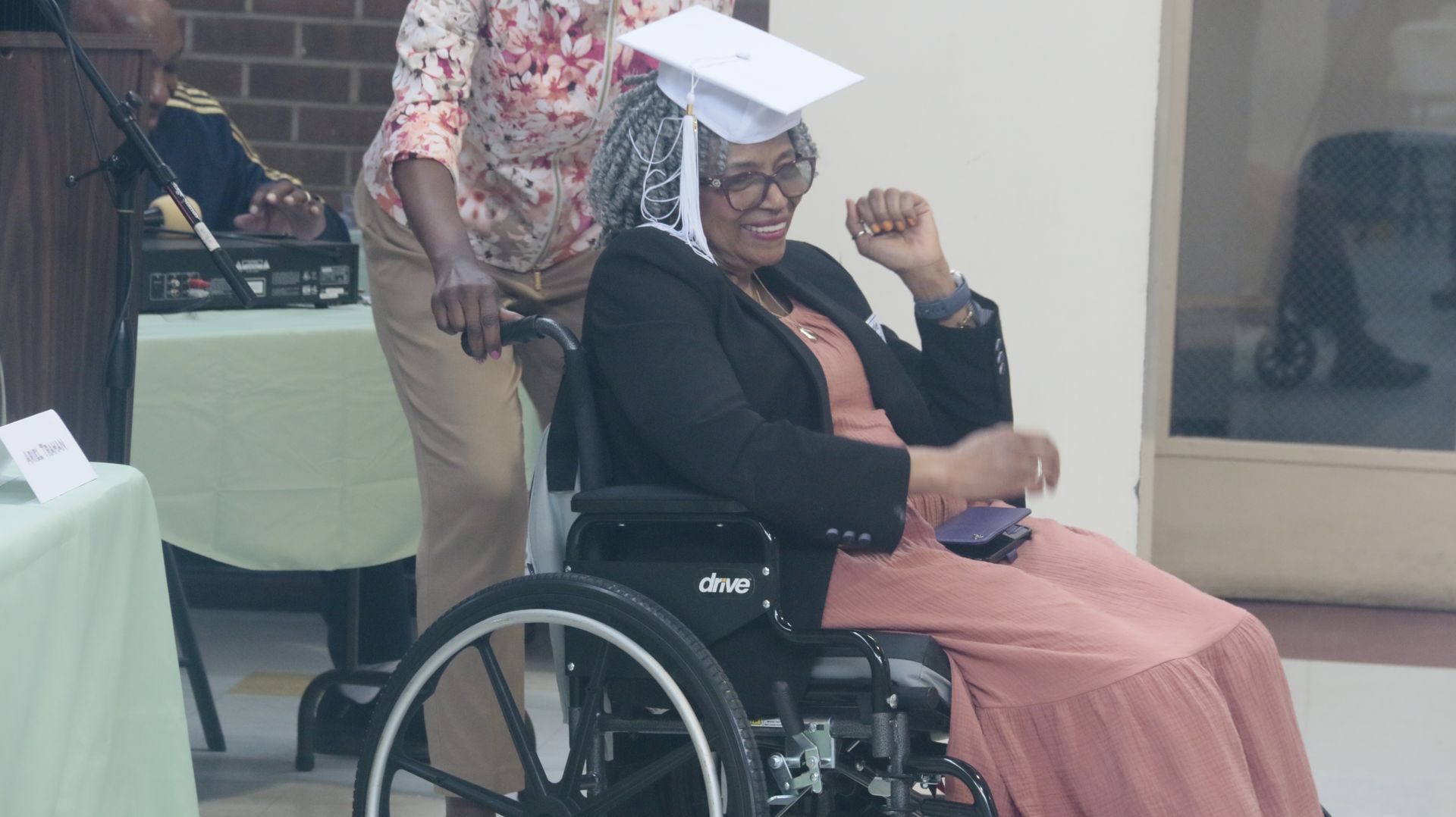 Person in graduation cap, wheelchair, orange dress, being assisted at an indoor event.