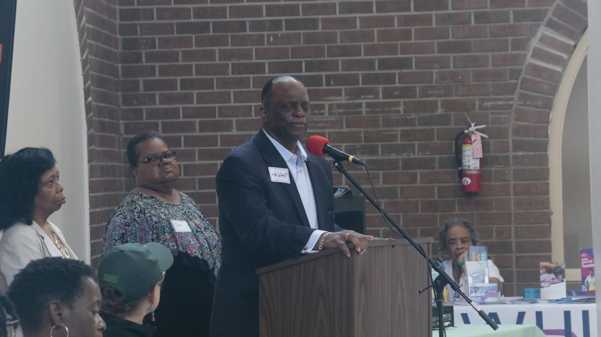 Man speaking at podium, standing in front of brick wall, other people behind him.