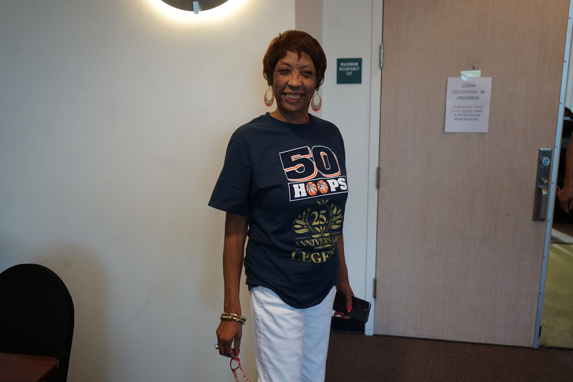 Woman in navy t-shirt and white pants smiles by a door; standing indoors.