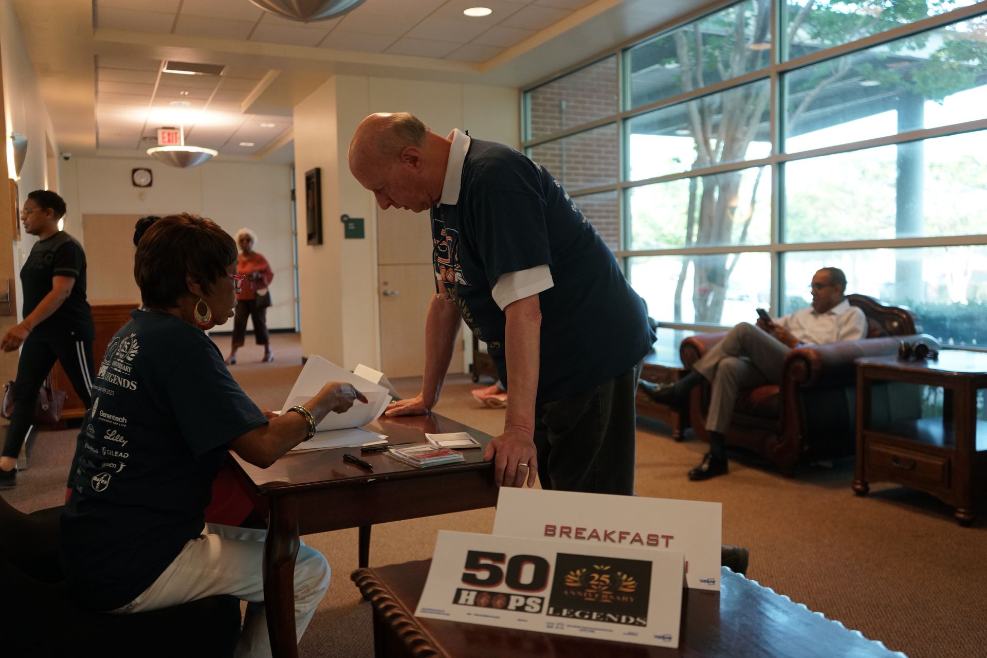 People at a table signing in for breakfast. A man leans over, a woman is sitting, and others are in the background.