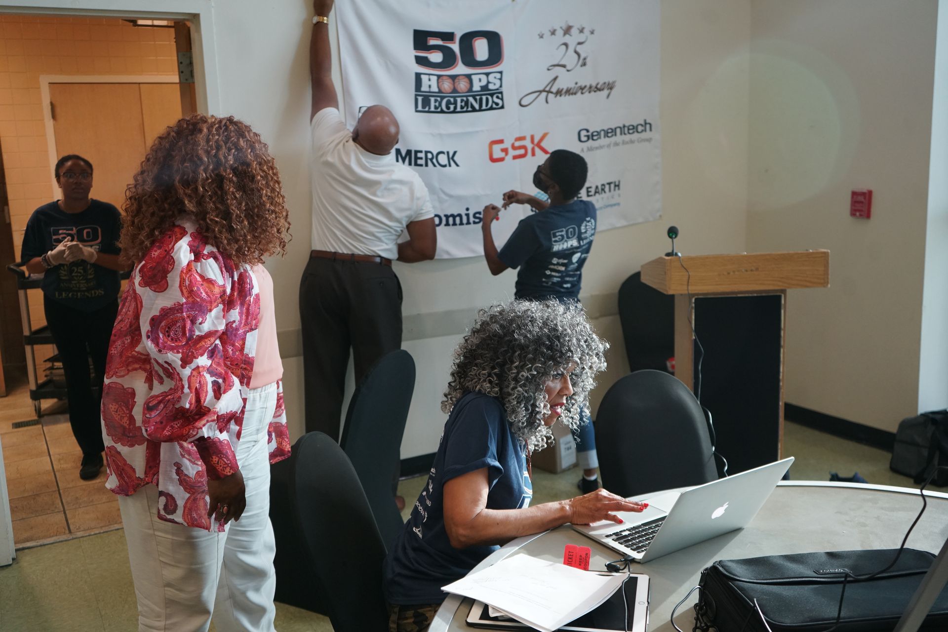People hanging a banner in a room. Others work at a laptop and stand by a doorway.