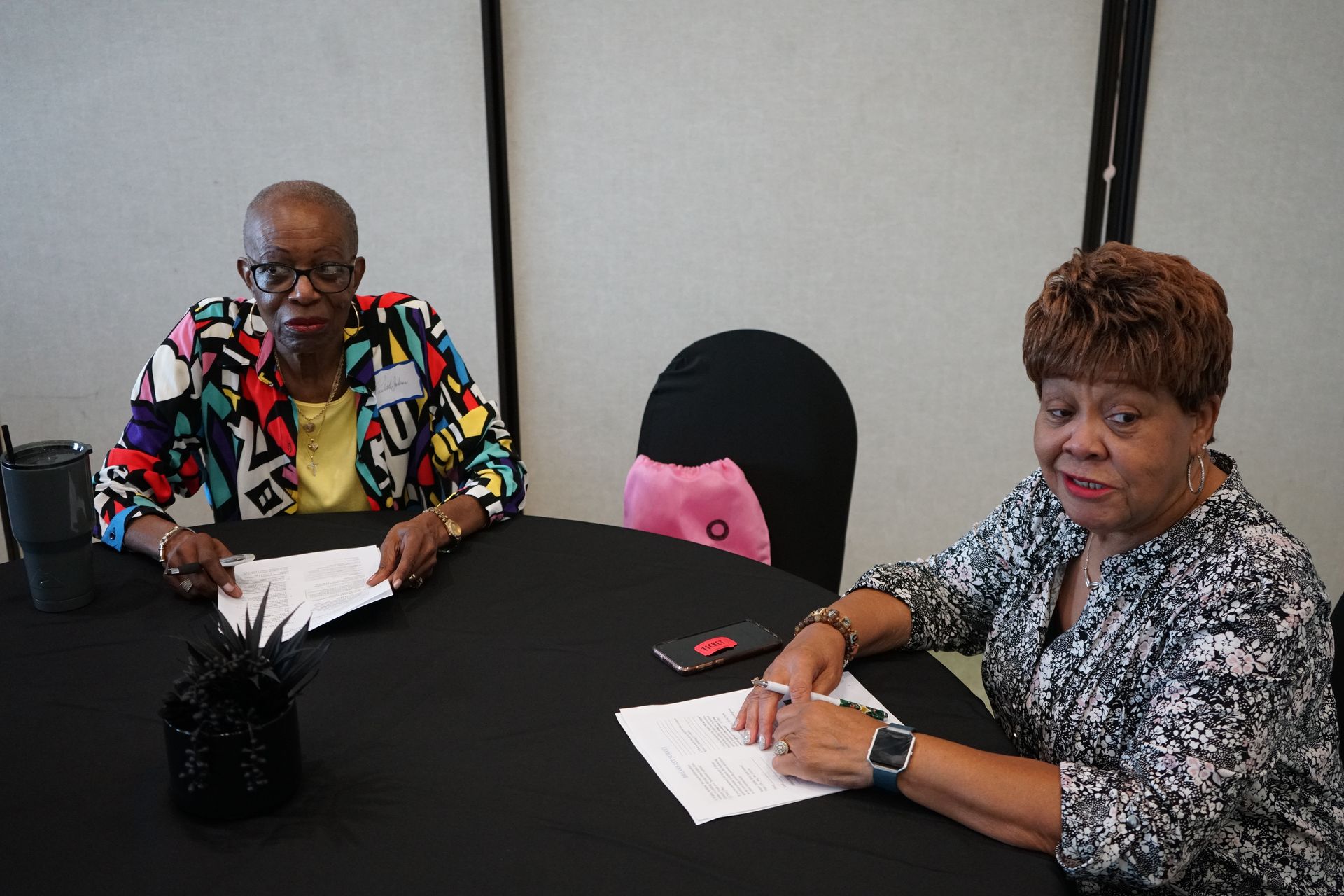 Two people sitting at a black table, reviewing documents. One wears a patterned jacket, the other a floral blouse.