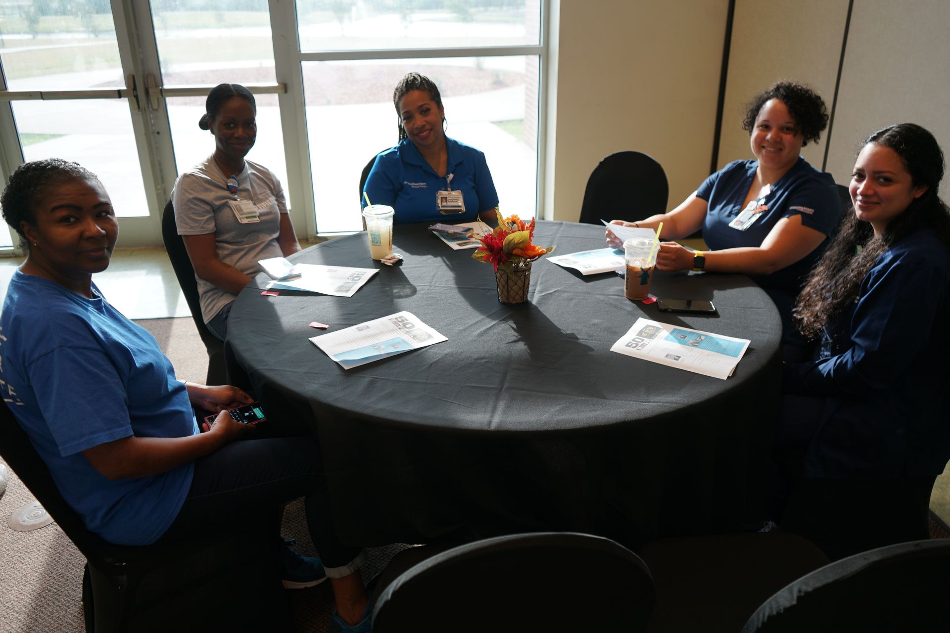 Five people in blue shirts seated around a black table, smiling. Window in the background.