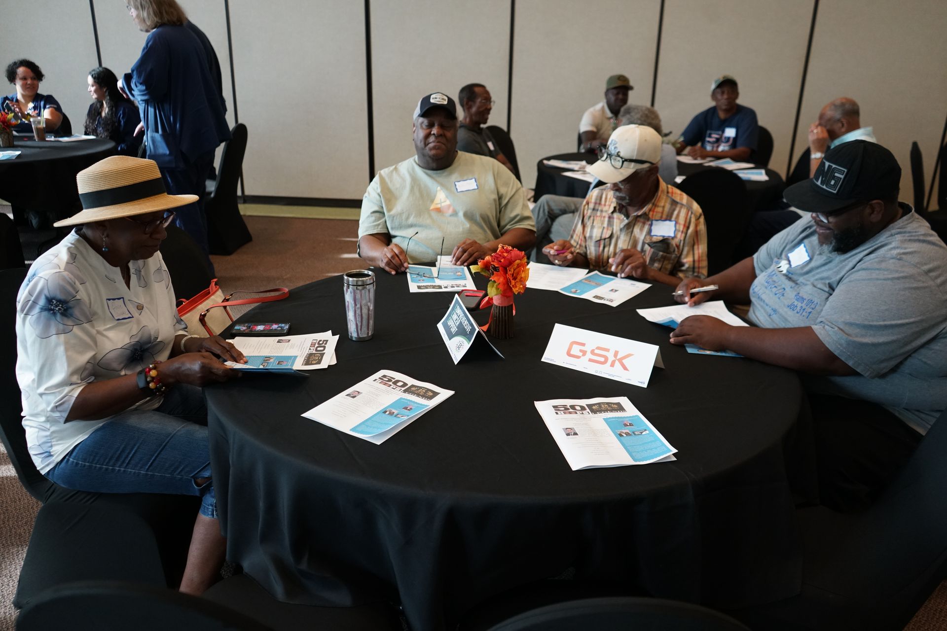 People seated at tables during a meeting. Papers, a flower arrangement, and name tags visible.
