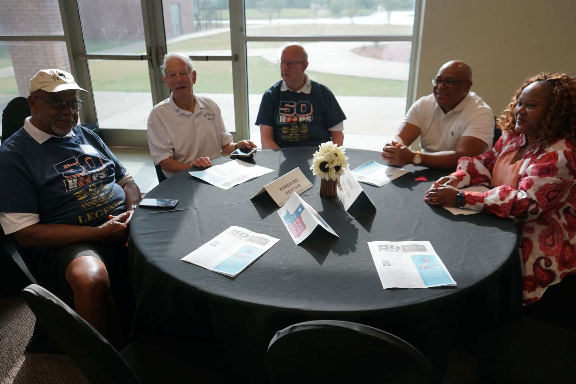 A diverse group of people seated around a table, documents and centerpieces are present.