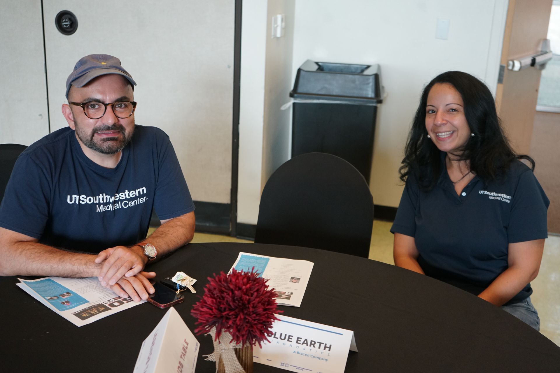 Two people seated at a table with papers, one wearing a cap and glasses, the other a polo shirt, indoors.