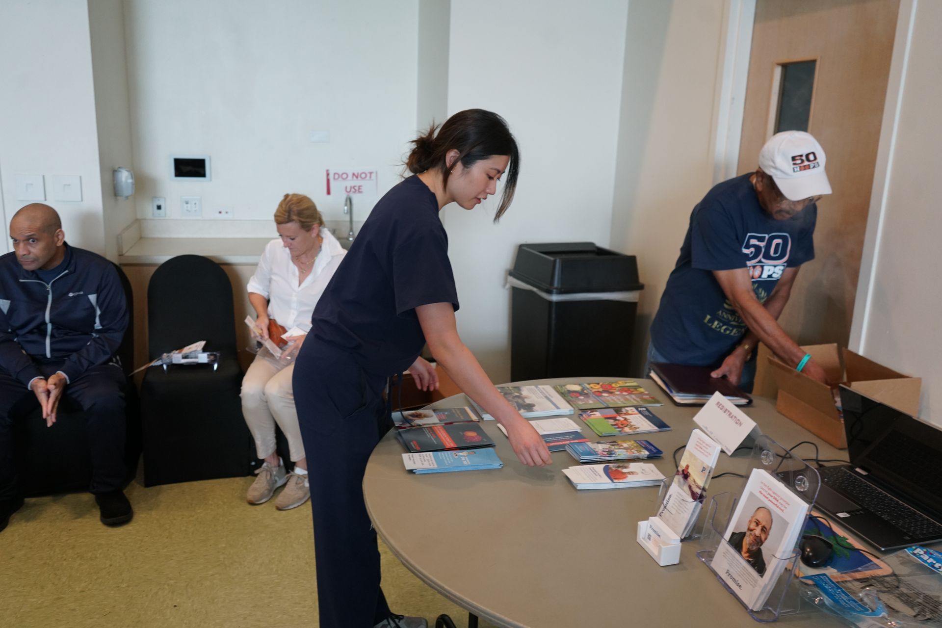 A woman arranging materials on a table, with two people seated nearby. A man is also preparing items. Indoors.