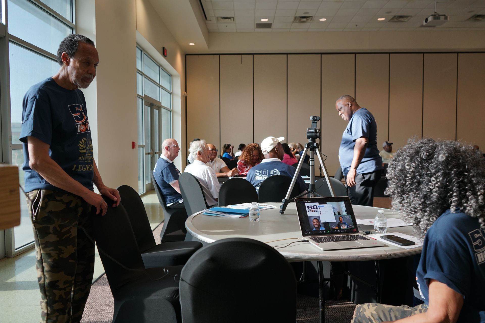 People in a conference room; some seated at round tables, others standing. A laptop sits on a table.