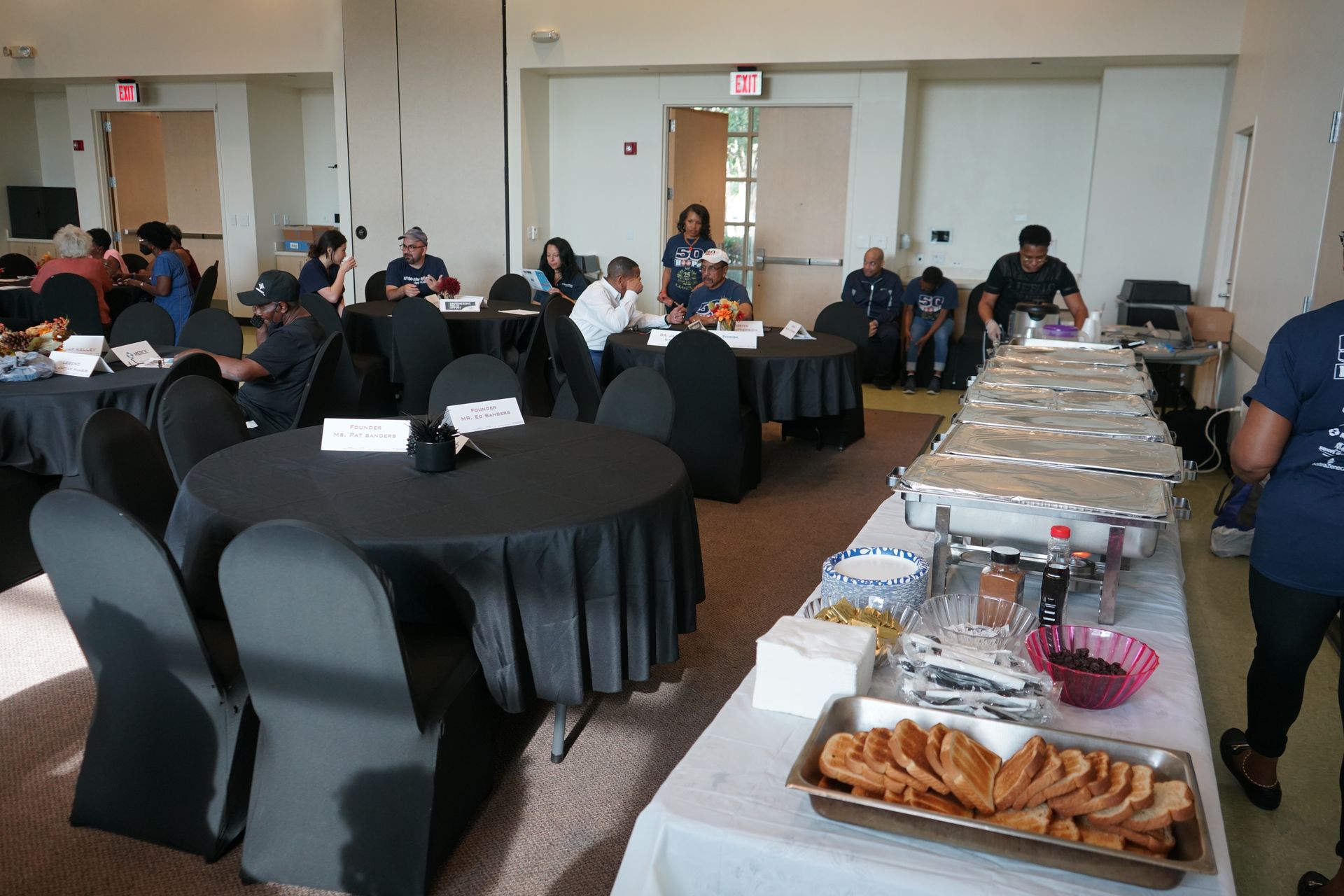 A room with round tables draped in black cloth, people seated, buffet table set up.