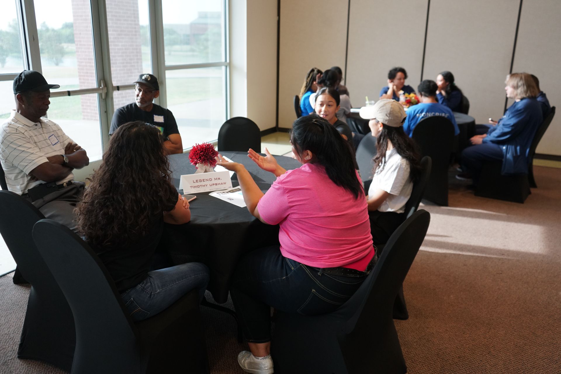 People sit around tables, engaged in conversation, in a bright room with a window.