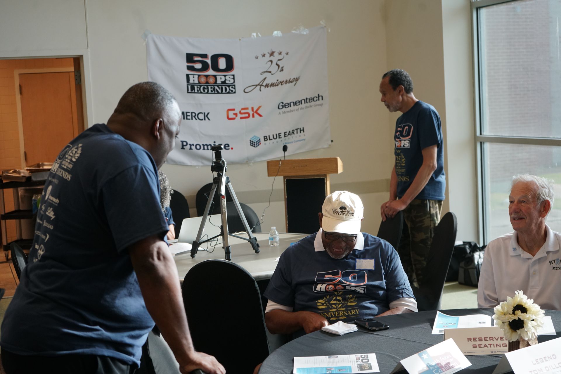 People at event with banner for 50th anniversary. They are interacting at tables in a room with a podium.