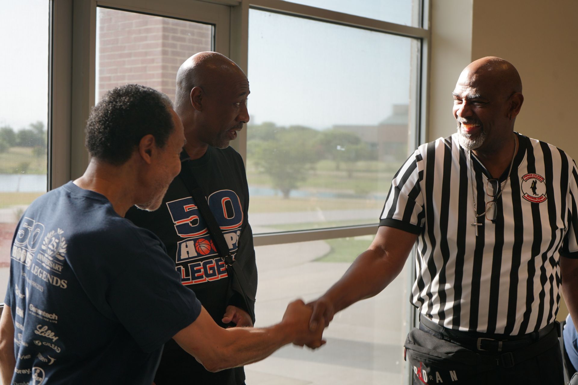 Three men shaking hands near a window. The man on the right wears a referee shirt.