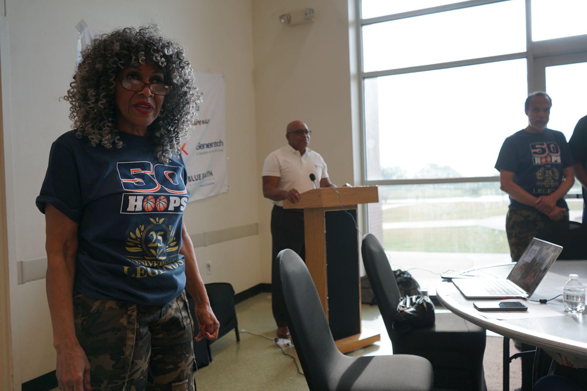 Woman in camo pants and a t-shirt stands near a table and a man at a podium; bright light from window.