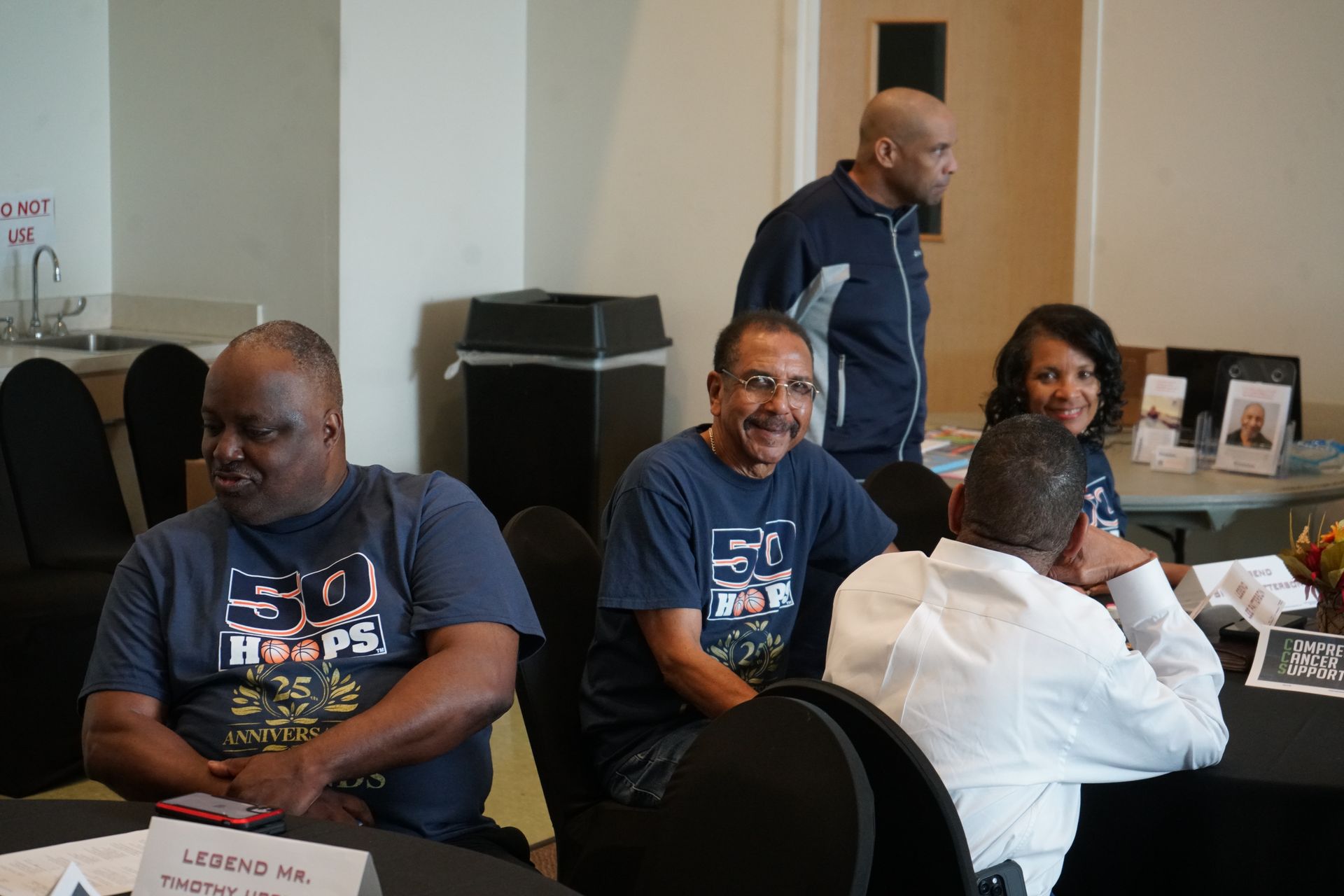 People seated at tables, smiling, in a well-lit room. Some wear blue shirts. One person standing in the background.