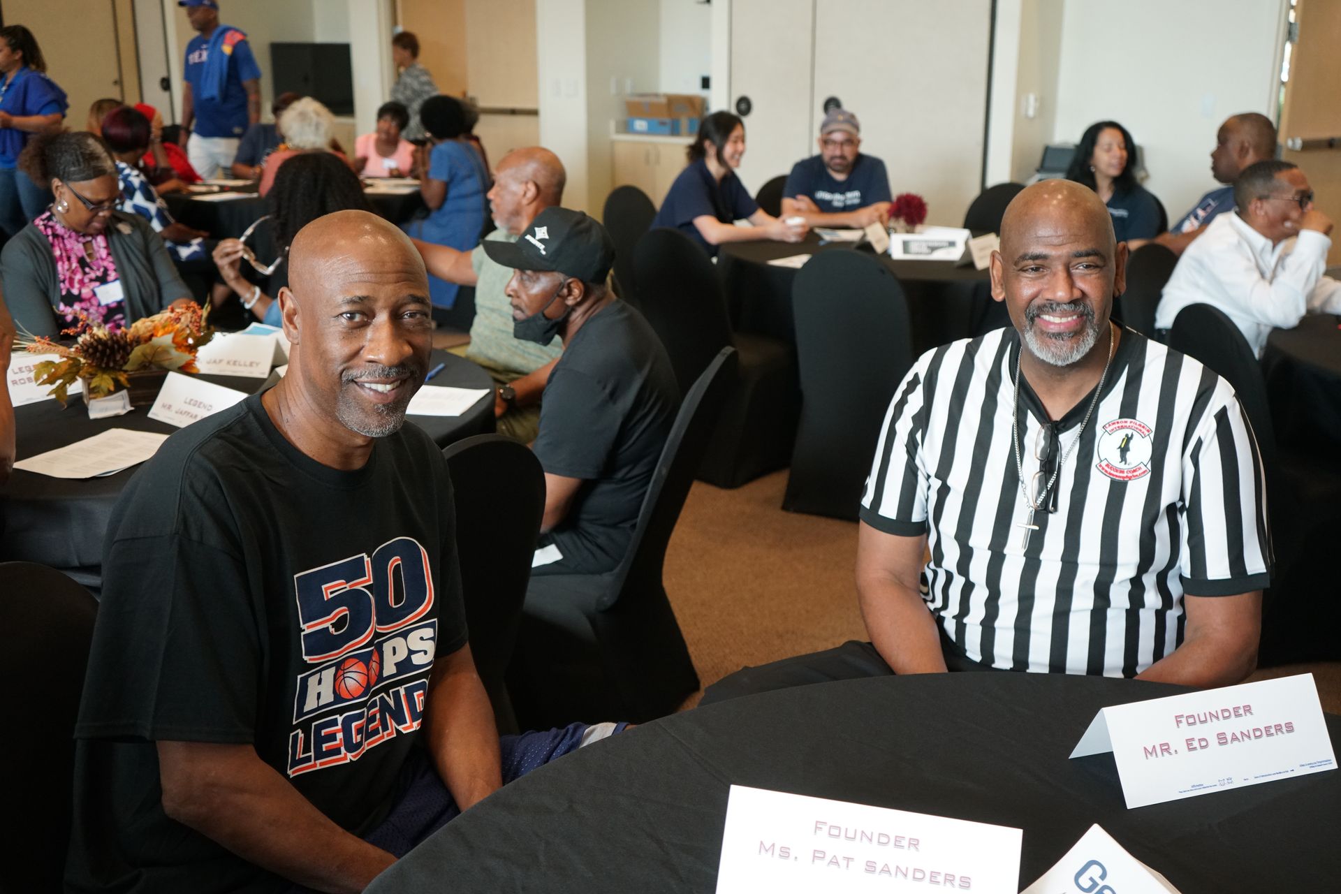 Two men at a table, one in referee stripes, others seated in a room, attending an event.