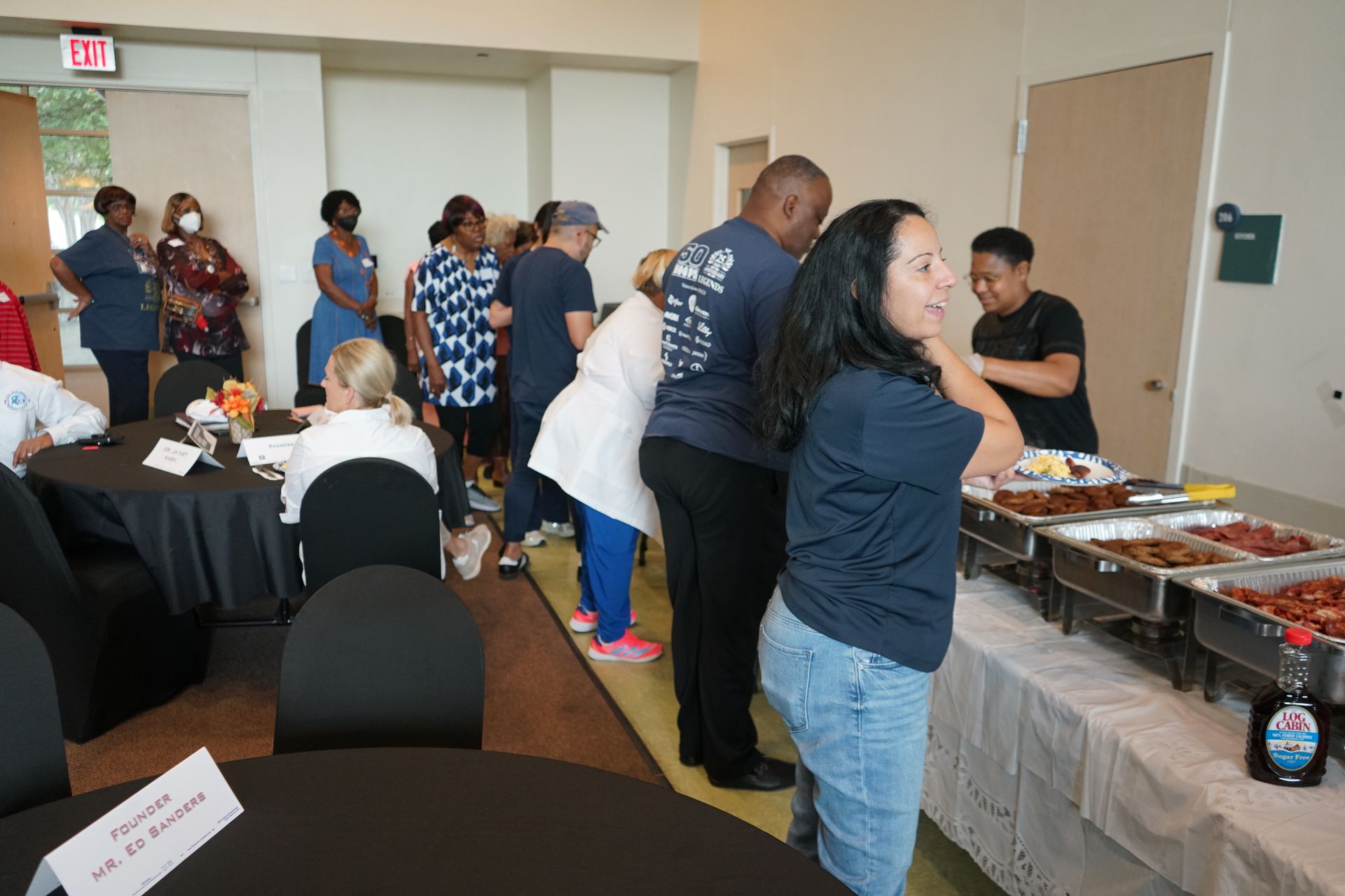 People line up at a buffet table with food. Other people seated at tables nearby.