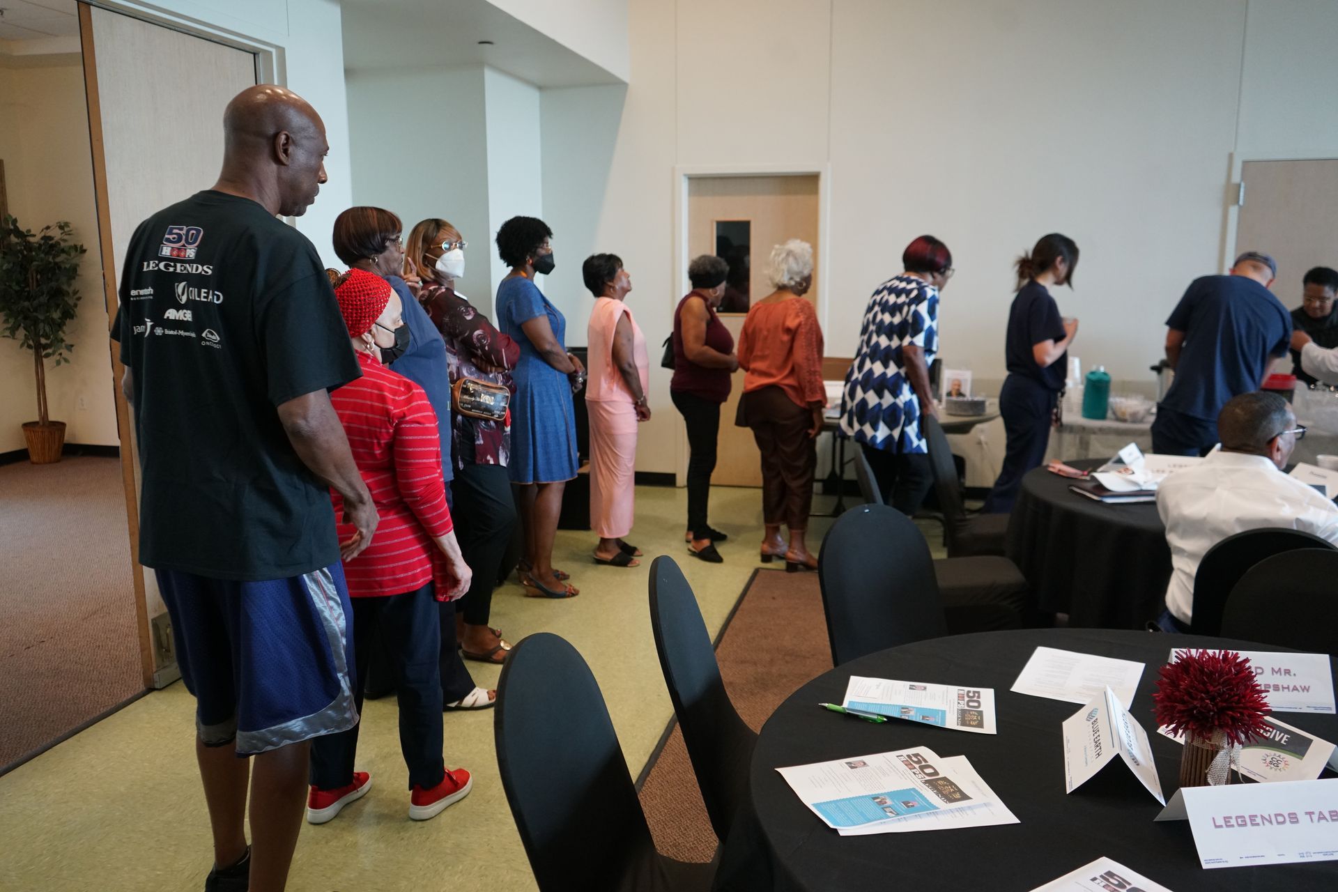 People line up near tables in a well-lit room. Some wear masks. Some are looking forward.